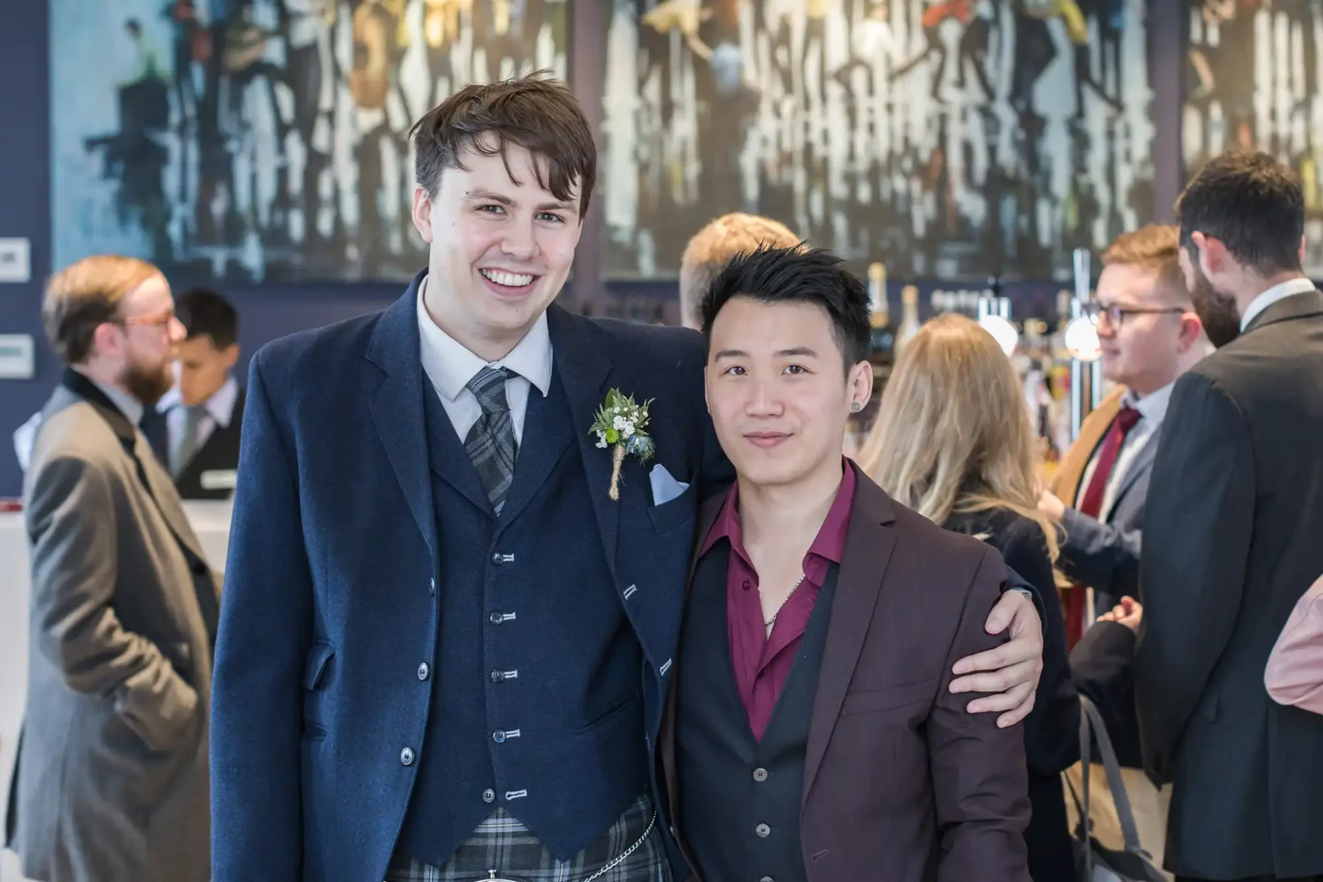 Two men smiling at a formal event, one wearing a traditional kilt and the other in a maroon jacket, with guests in the background.