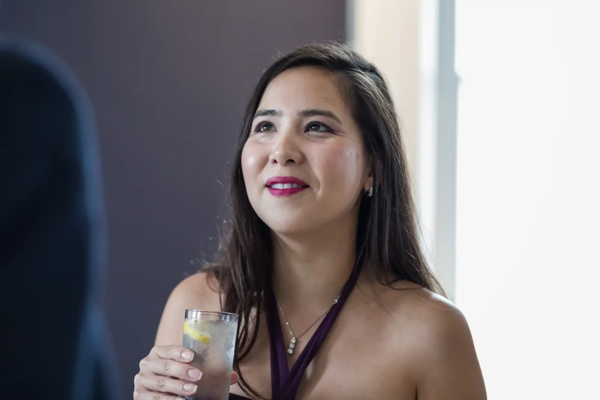 Woman holding a glass, smiling and looking up, engaged in conversation at a social event.