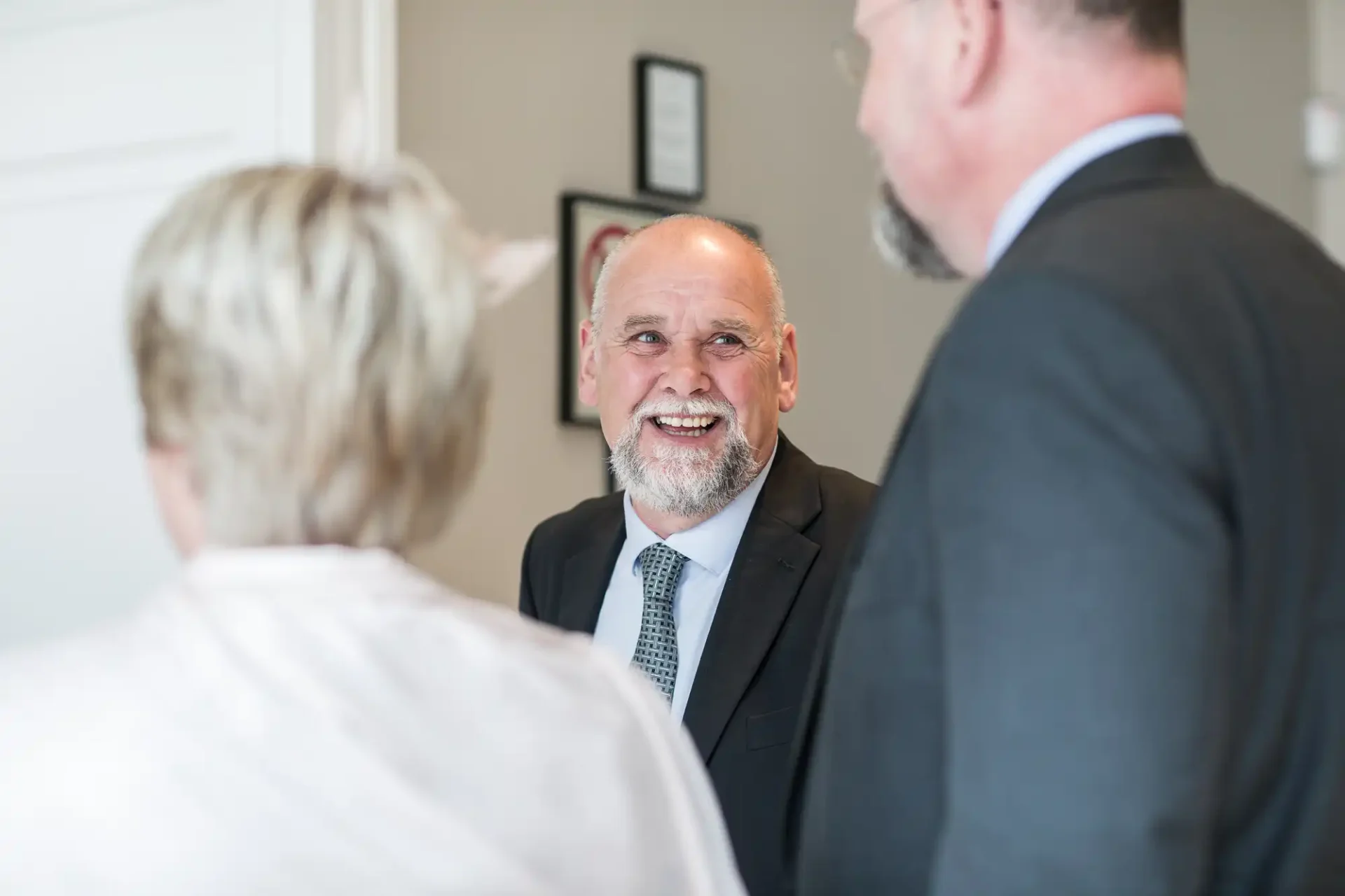 A smiling bald man with a beard, wearing a suit, conversing with two people whose backs are to the camera in a room with framed pictures on the wall.