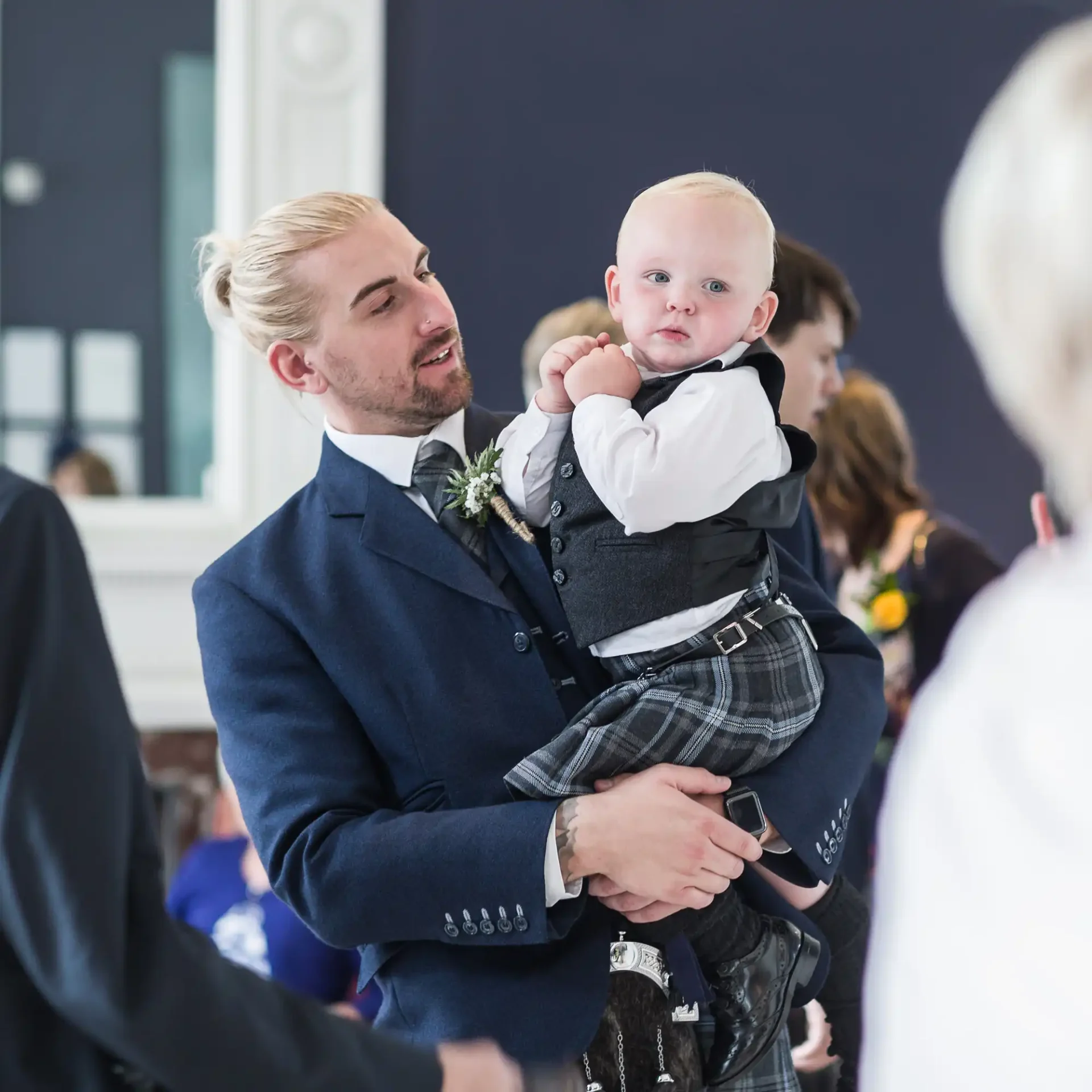 A man in a blue suit holding a baby dressed in a kilt and vest at a formal gathering.