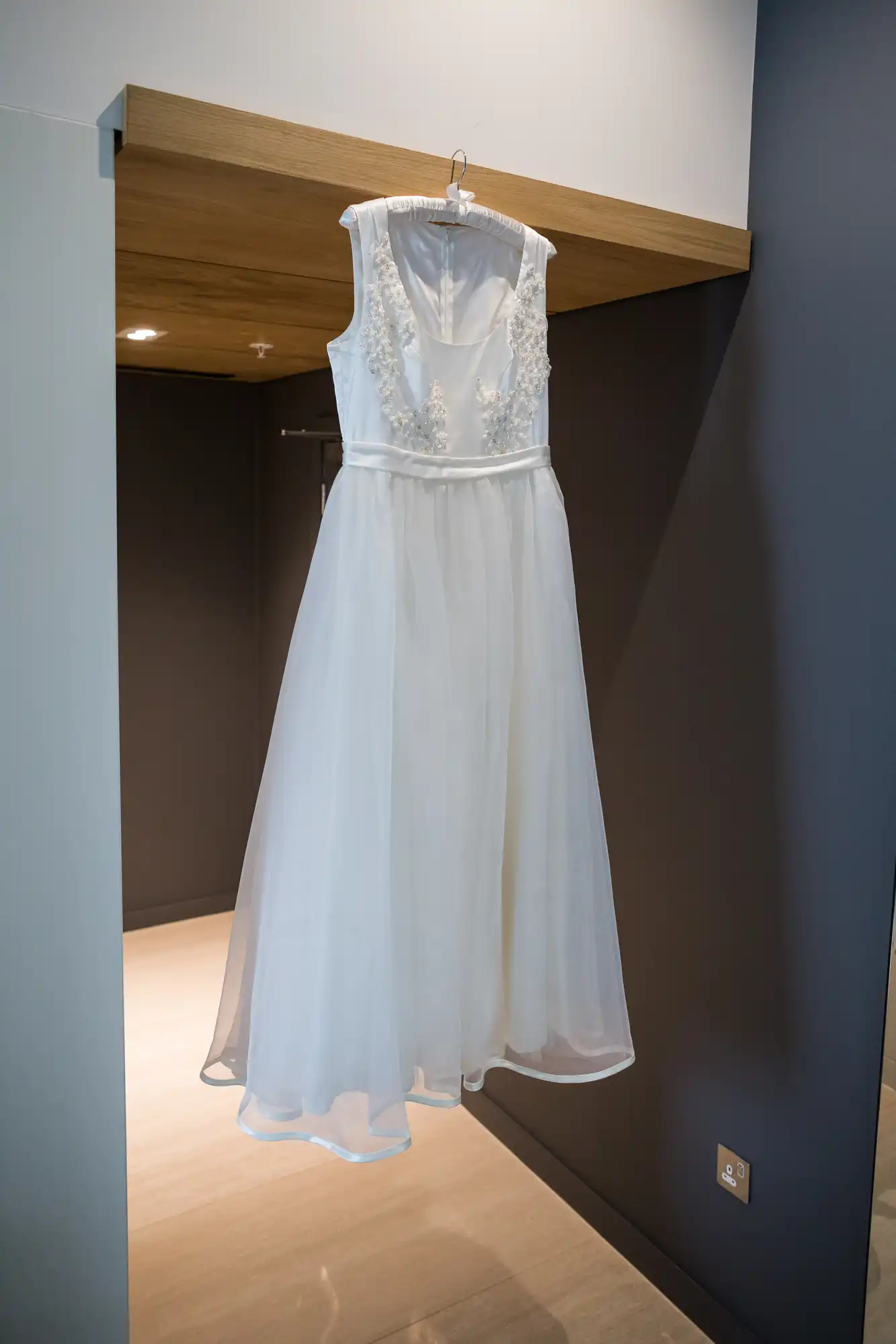 A white wedding dress with lace details hanging on a hanger against a dark blue wall with wood paneling above.