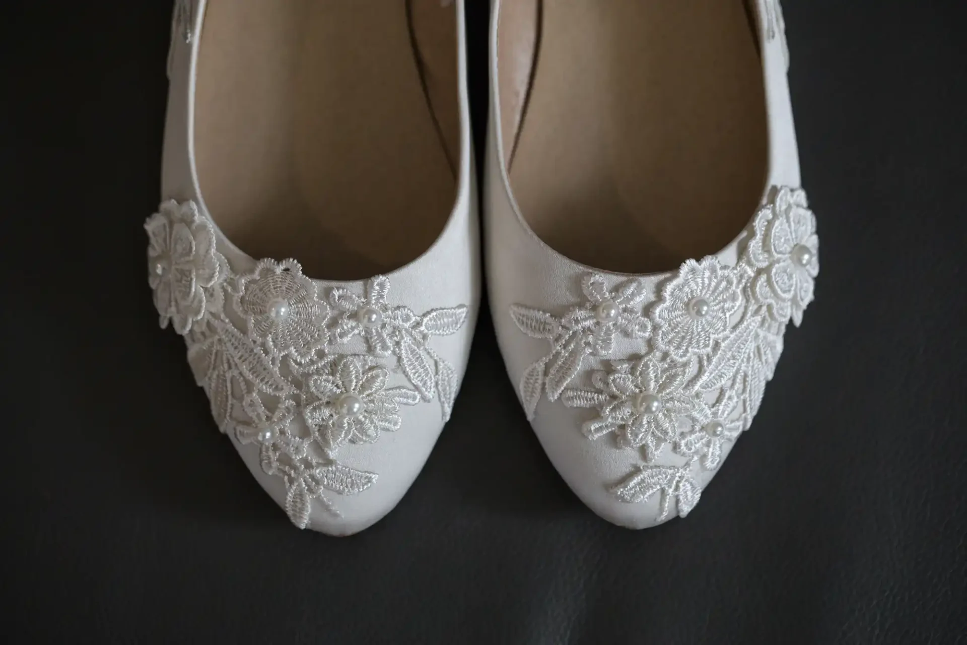 A pair of white bridal shoes with lace and bead detailing, displayed on a dark background.