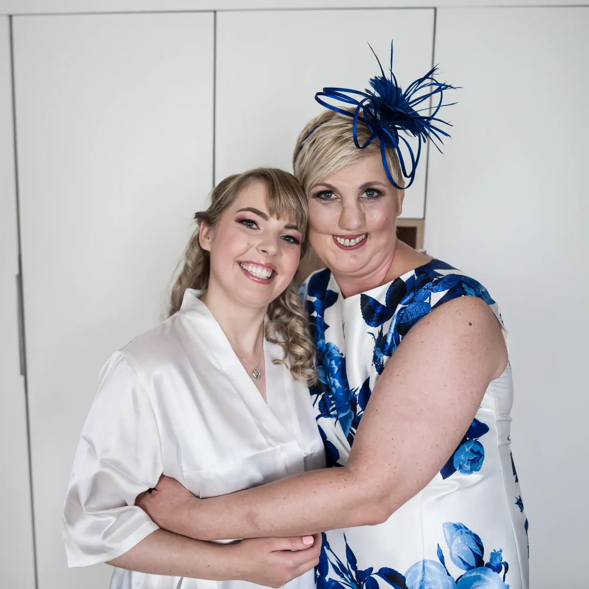 Two women smiling and embracing, one in a white robe and the other in a blue floral dress with a matching hat.