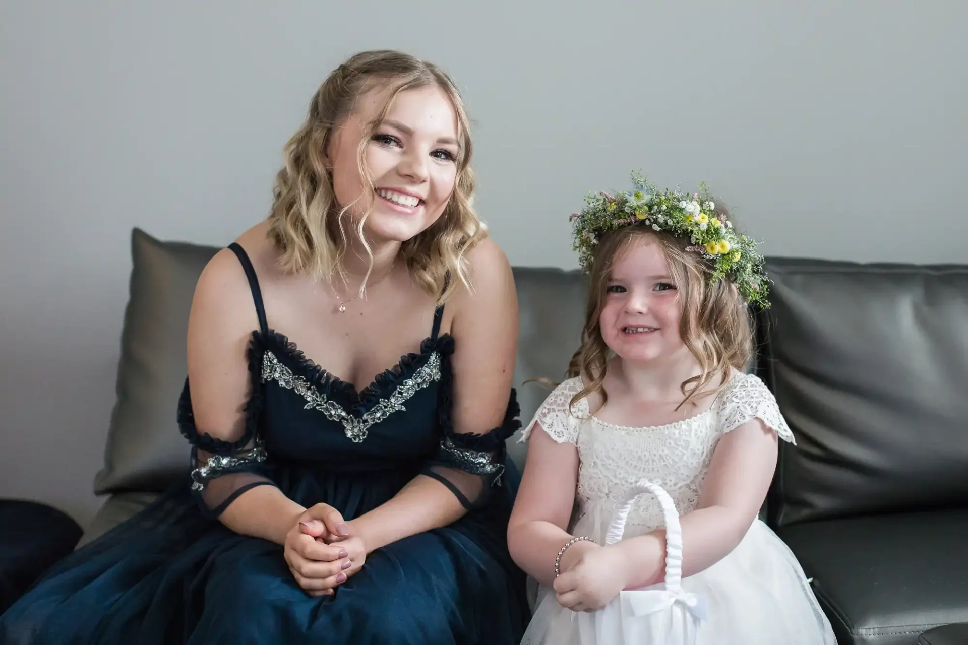 Two smiling females, an adult and a child, sitting on a couch. the adult wears a dark dress and the child dons a white dress with a floral headband.