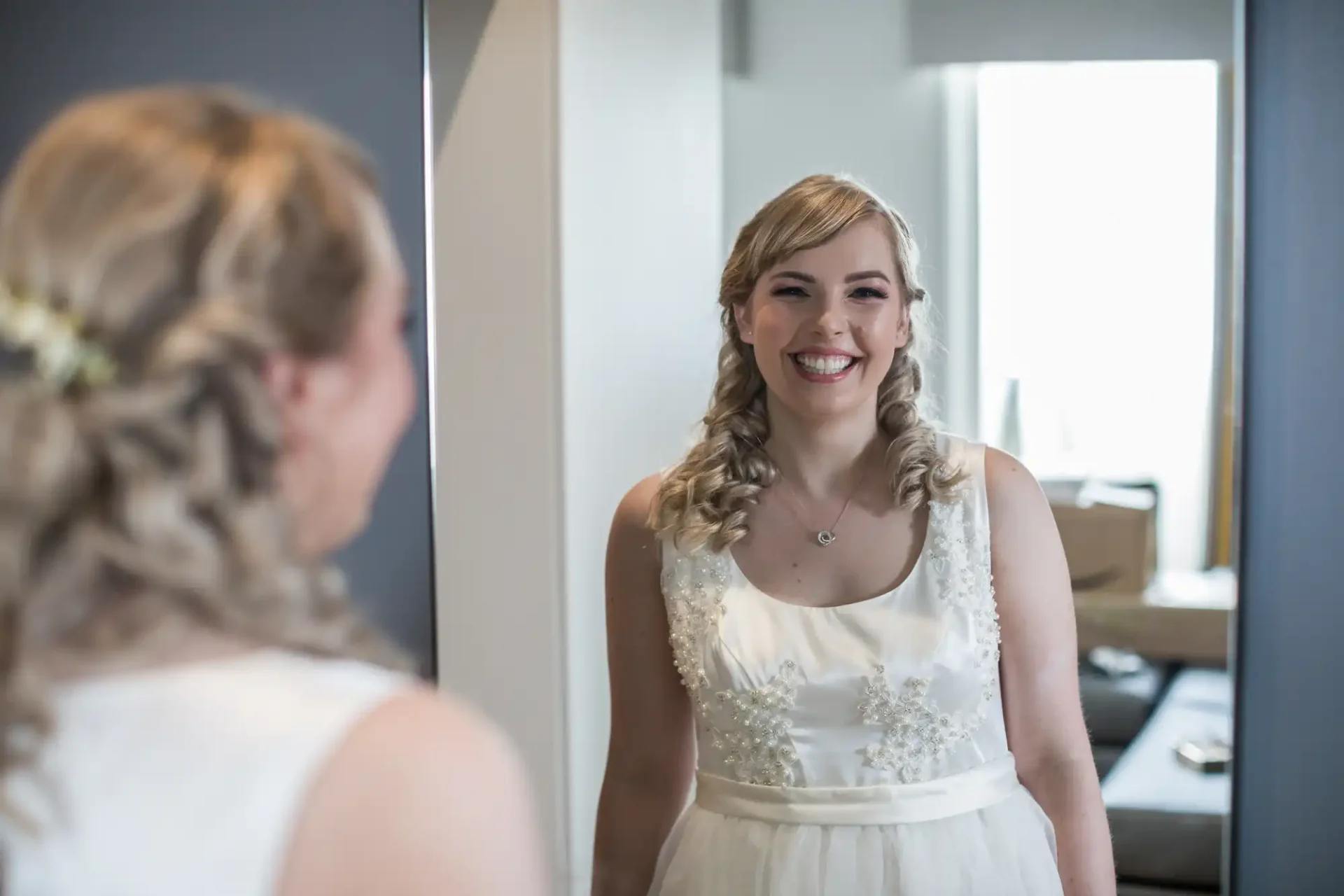 A woman in a white wedding dress smiling at her reflection in a mirror, with a styled braid and floral hair accessory.