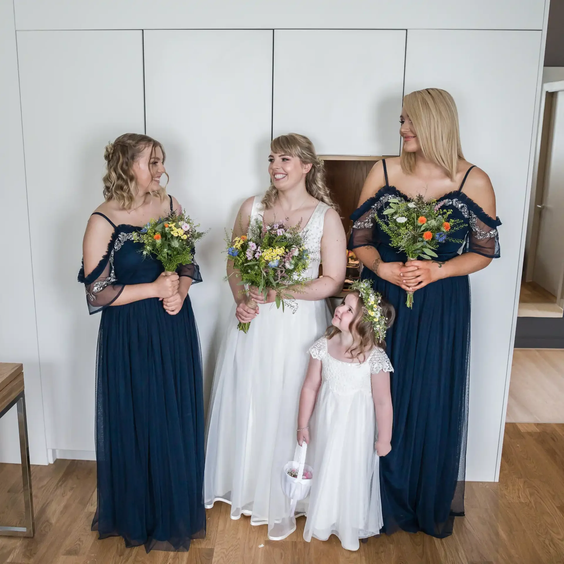 A bride in a white dress and three bridesmaids in navy dresses, holding bouquets, smiling and interacting joyfully indoors.