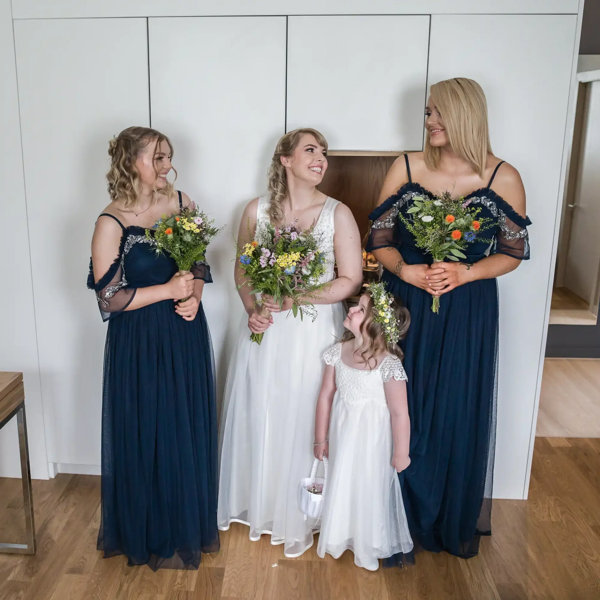 A bride in a white dress and three bridesmaids in navy dresses hold bouquets in a room, smiling and looking at each other.