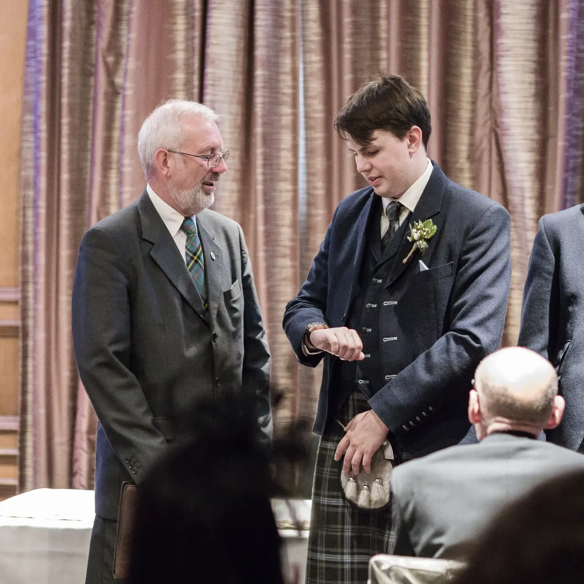 Two men in formal wear, one in a suit and the other in a kilt, smiling and talking at a wedding reception.