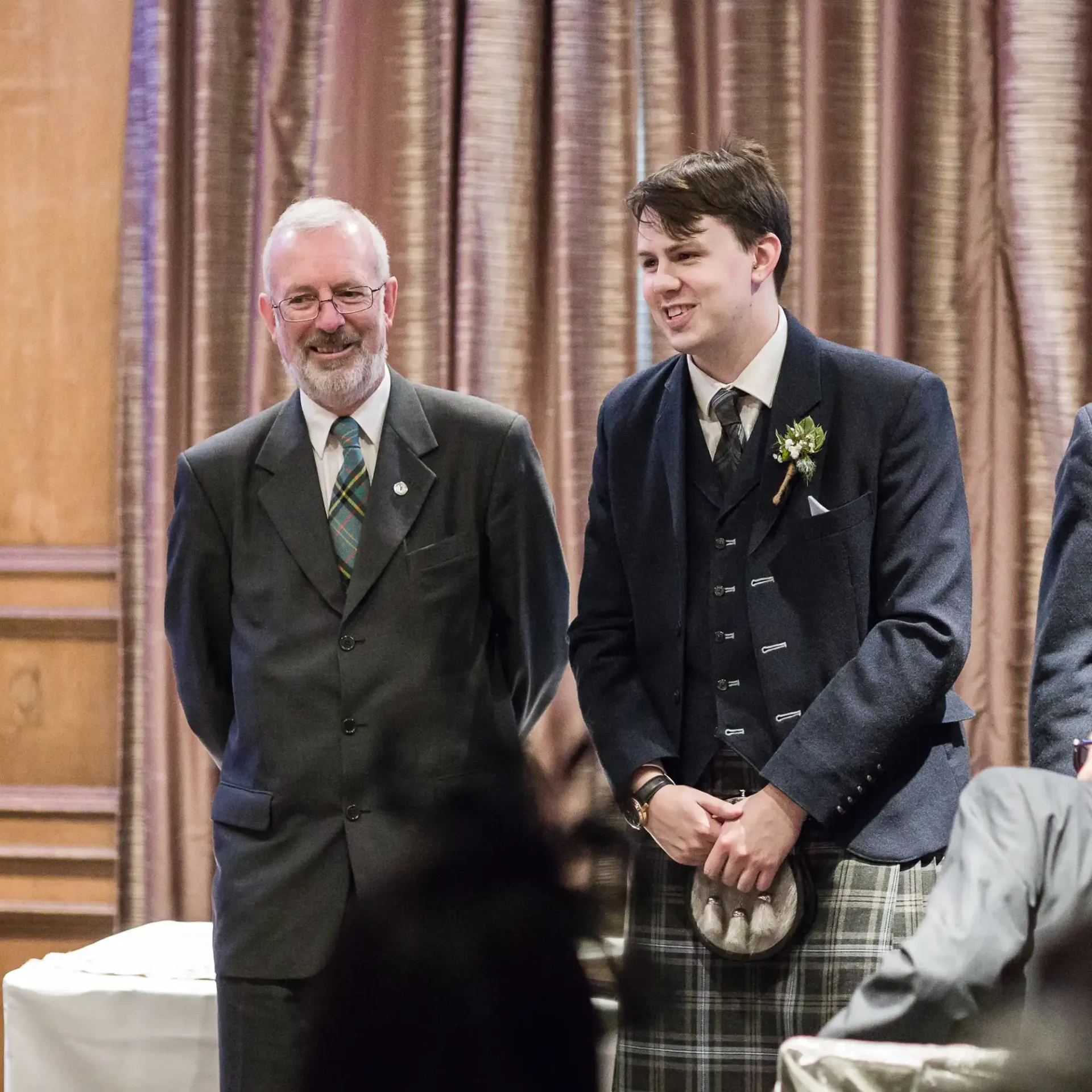 Two men smiling in a ceremonial hall, one wearing a suit and the other in a kilt, standing near a table with blurred guests in the foreground.