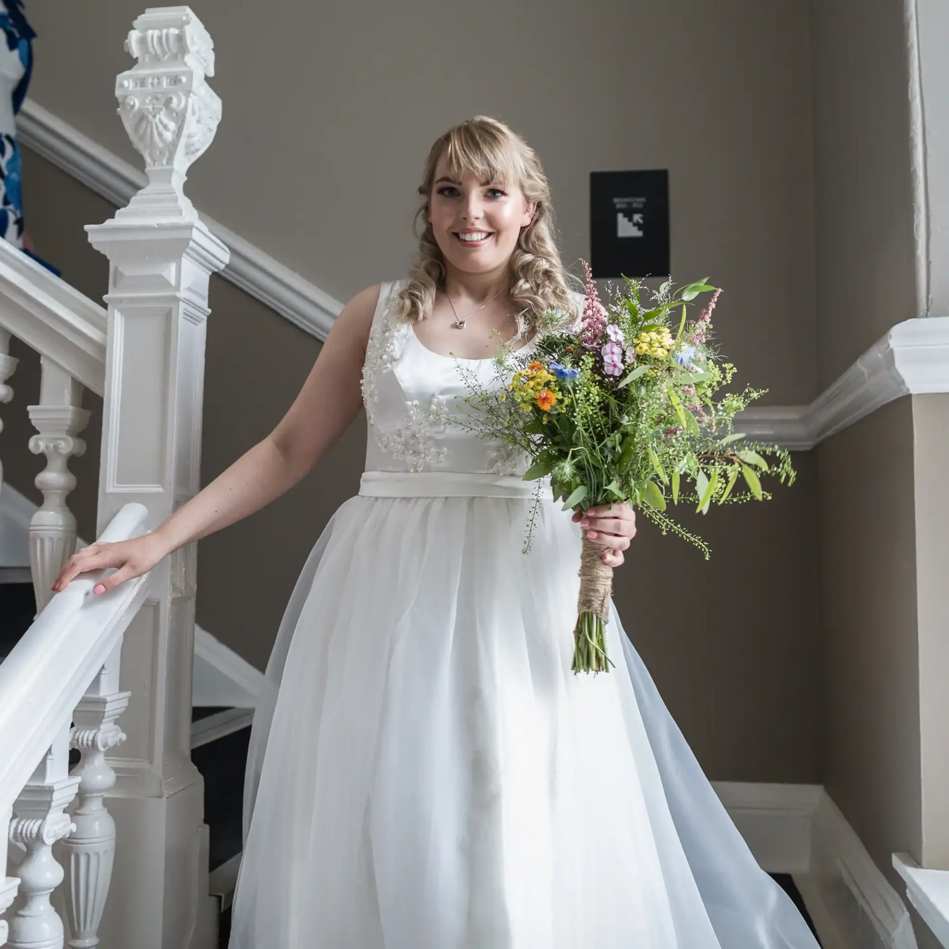 A bride in a white dress smiling, holding a bouquet of wildflowers while standing on a staircase with intricately designed railings.