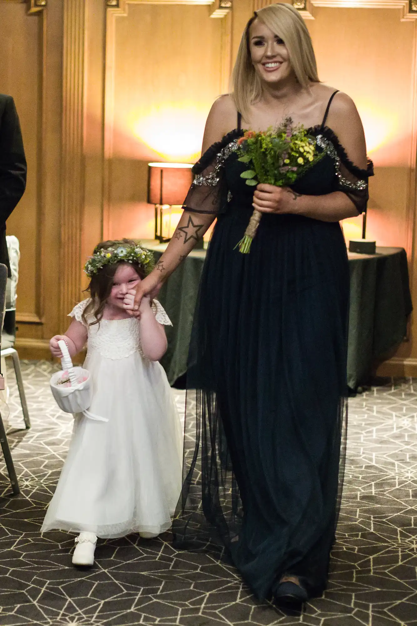 A woman in a navy dress holds a bouquet and the hand of a small girl in a white dress wearing a floral headband, both smiling as they walk.