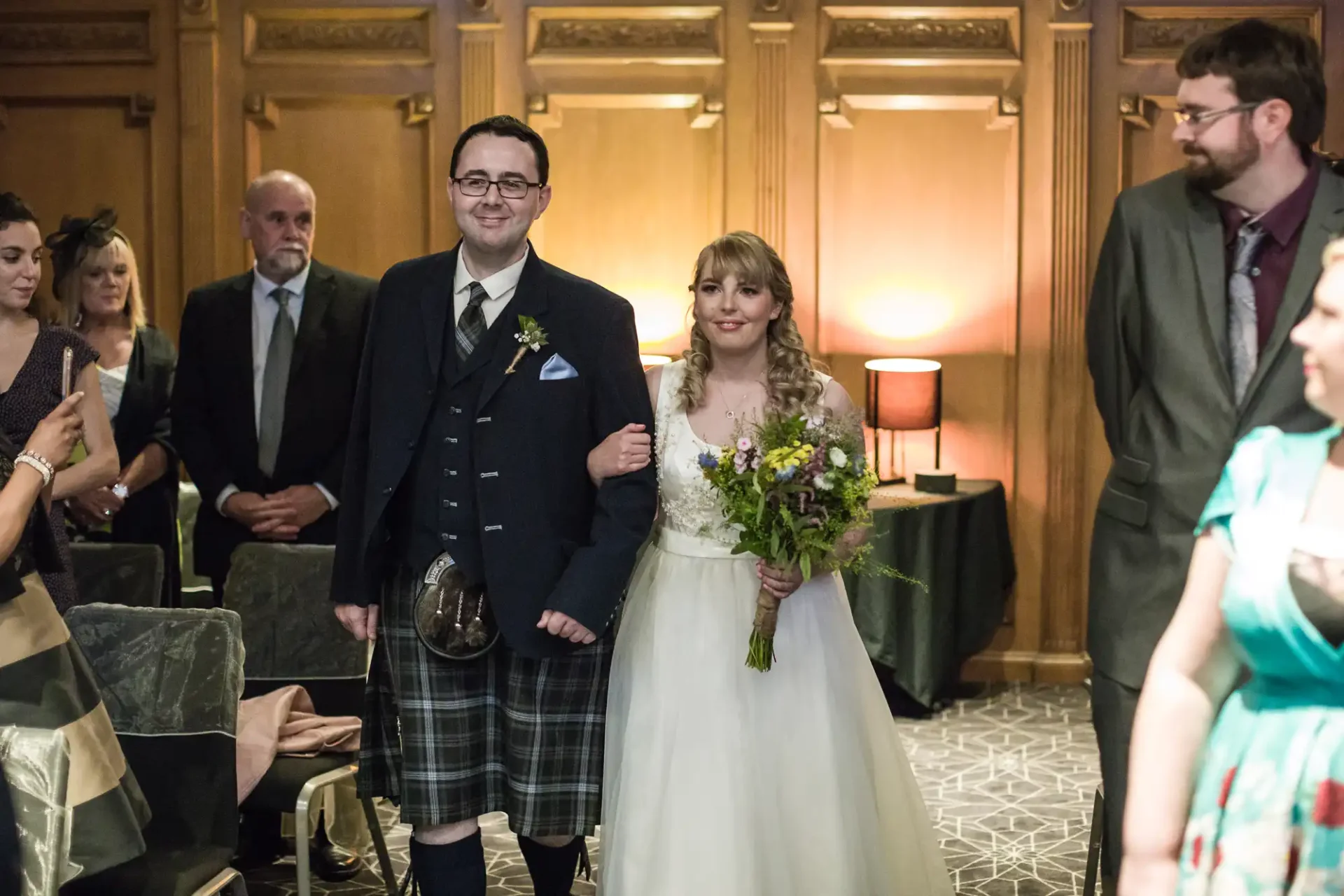 A bride and groom smile as they walk down the aisle, the groom wearing a kilt and the bride in a white dress, holding a bouquet, with guests watching.