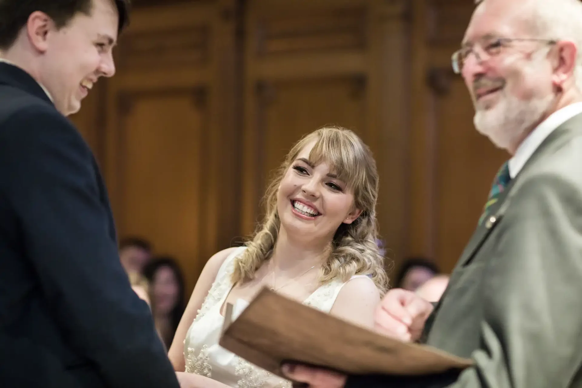 Bride and groom smiling at each other during their wedding ceremony, with an officiant holding a book in the foreground.