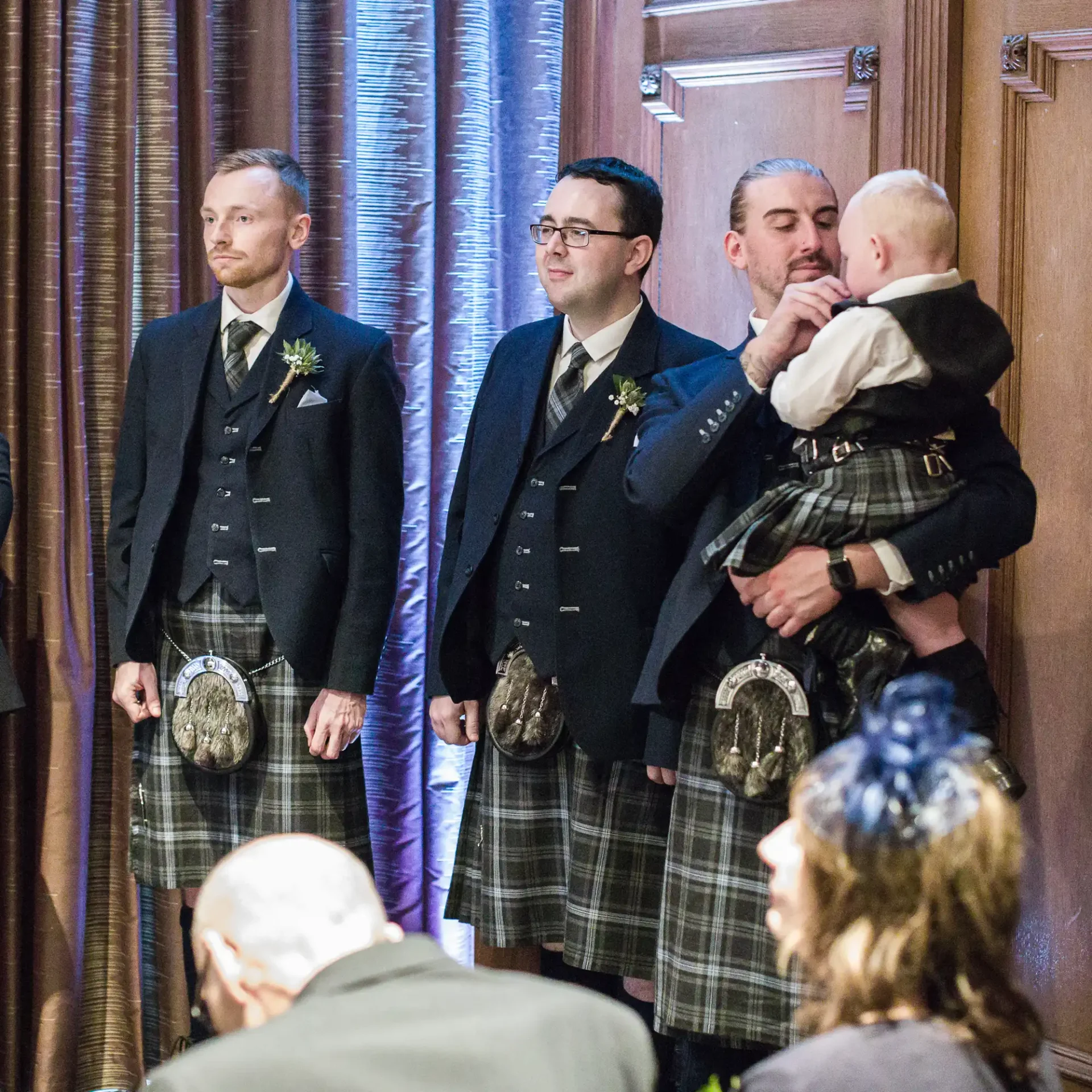 Three men in traditional scottish kilts at a formal event, one holding a baby, standing indoors with spectators in the background.