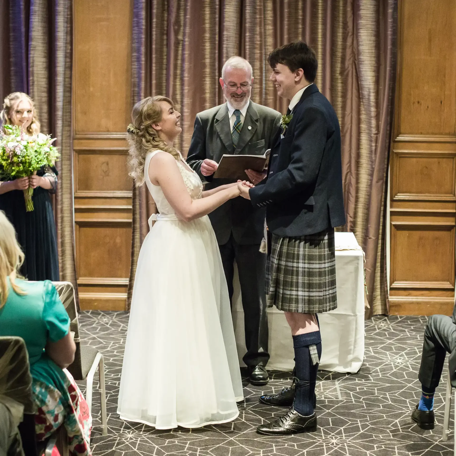 A bride and groom exchange vows, smiling joyfully at each other, while an officiant oversees the ceremony in a hall with guests watching.