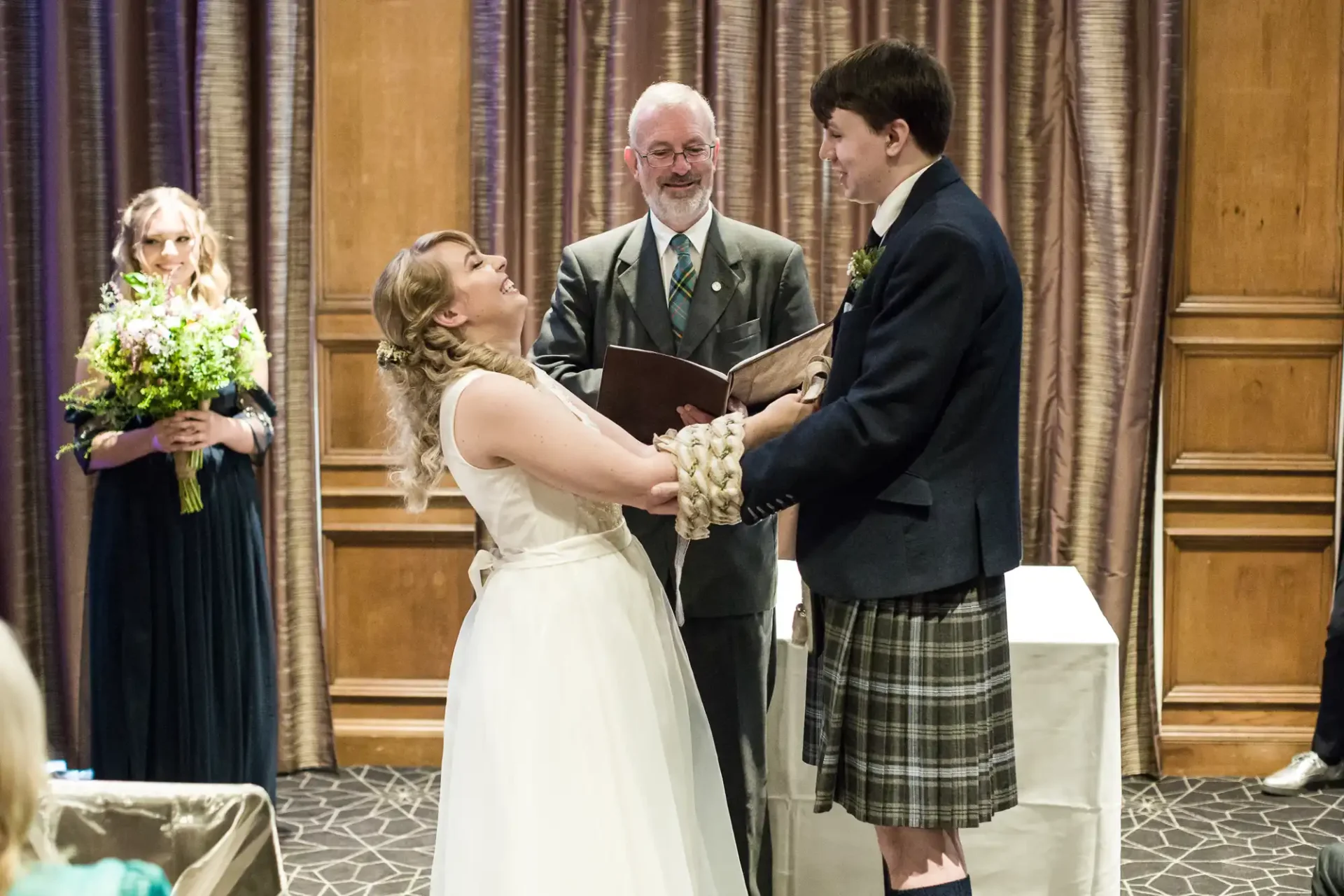 A bride and groom, wearing traditional scottish attire, hold hands during their wedding ceremony, with an officiant smiling behind them and a bridesmaid in the background.