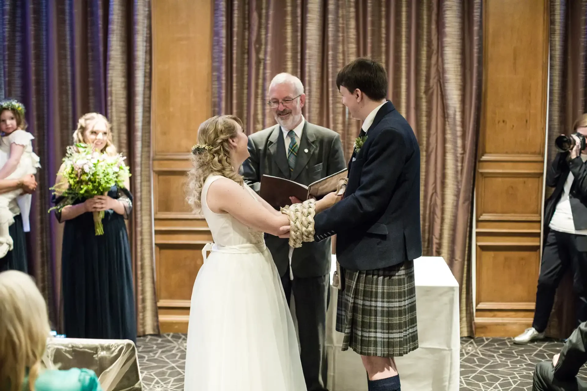 A bride in a white dress and a groom in a kilt exchanging vows at a wedding ceremony, with a minister overseeing and guests watching.
