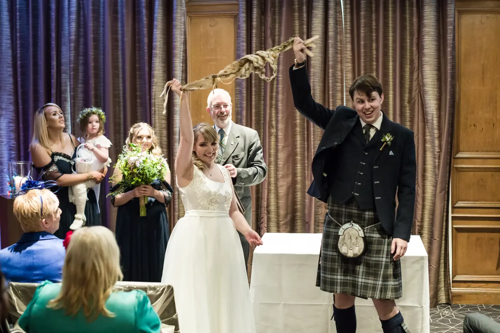 A bride and groom joyfully cut their wedding cake as guests watch, with the groom wearing a traditional kilt and the bride in a white dress.