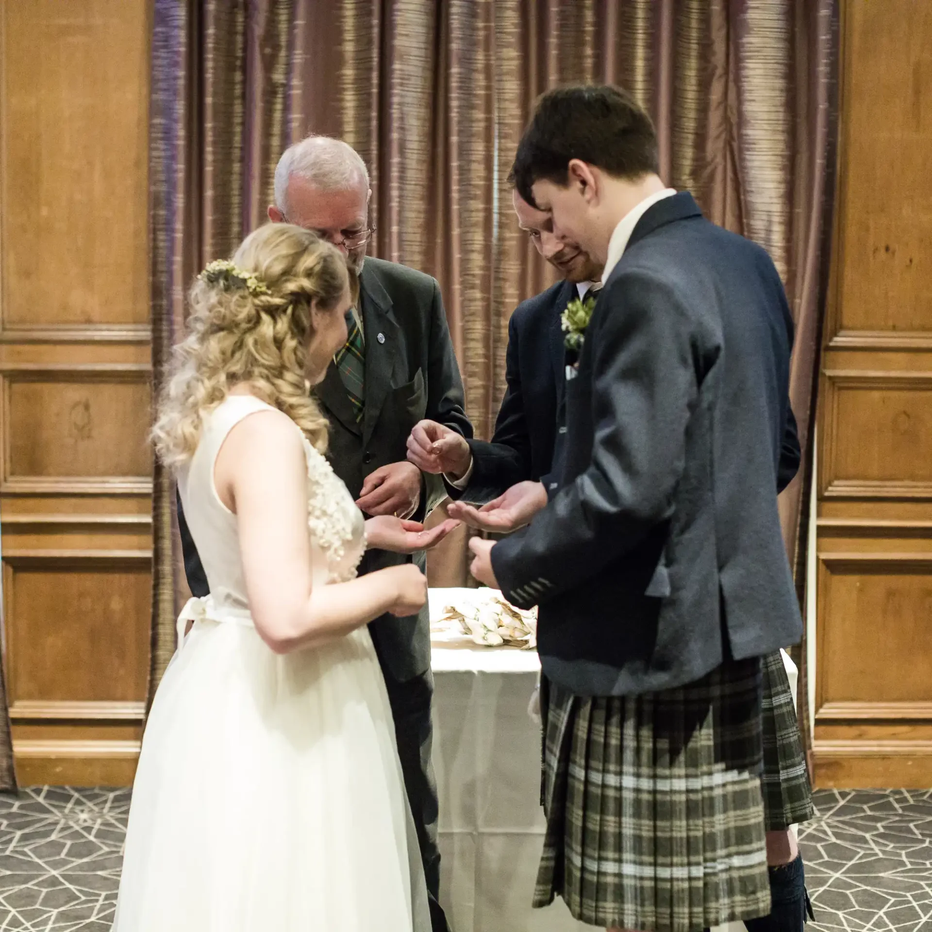 A bride and groom cut their wedding cake as an older man watches, with the groom wearing a kilt and the bride in a white dress.