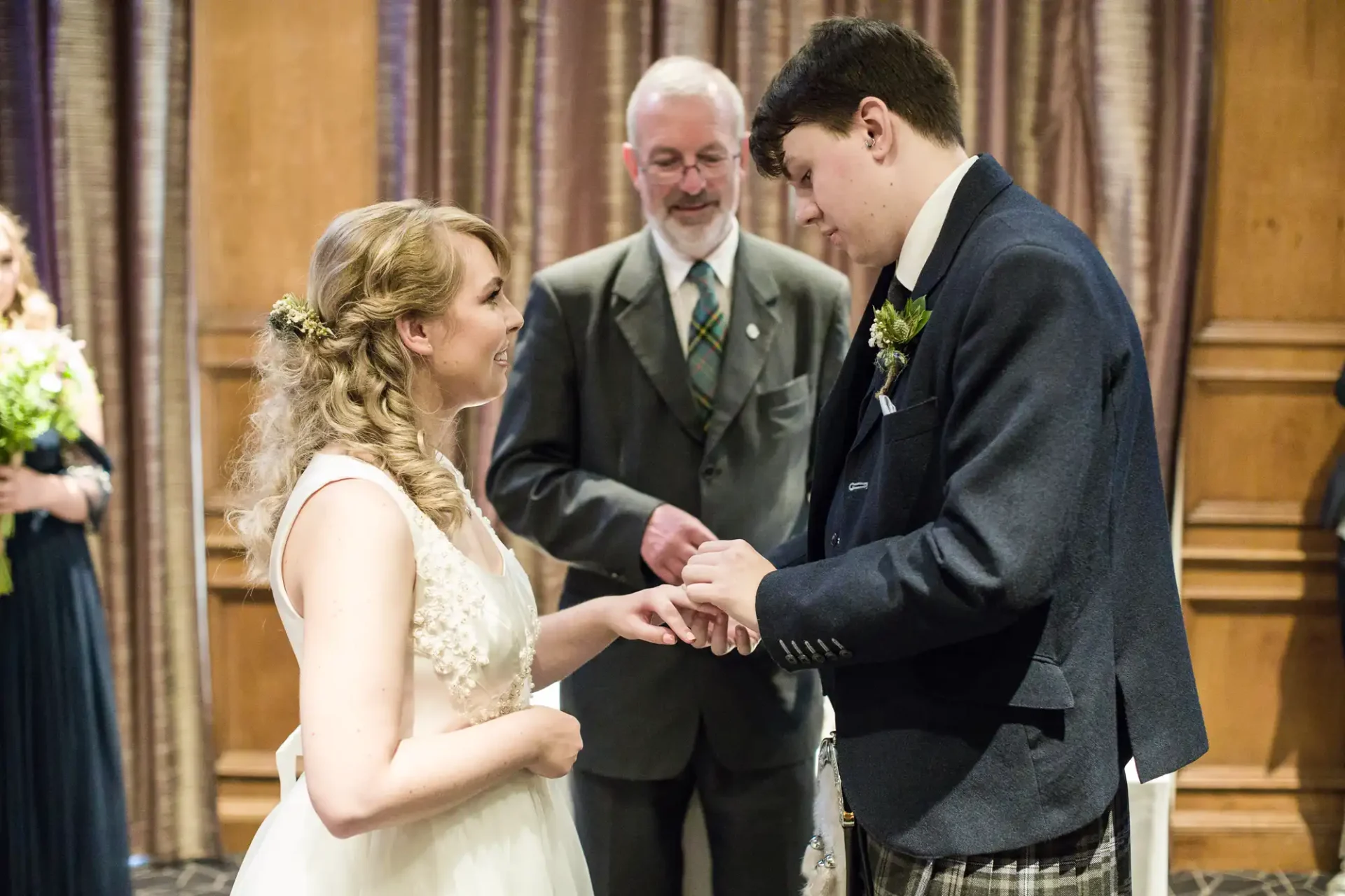 A bride and groom exchange rings at their wedding ceremony, with an officiant and bridesmaid in the background.