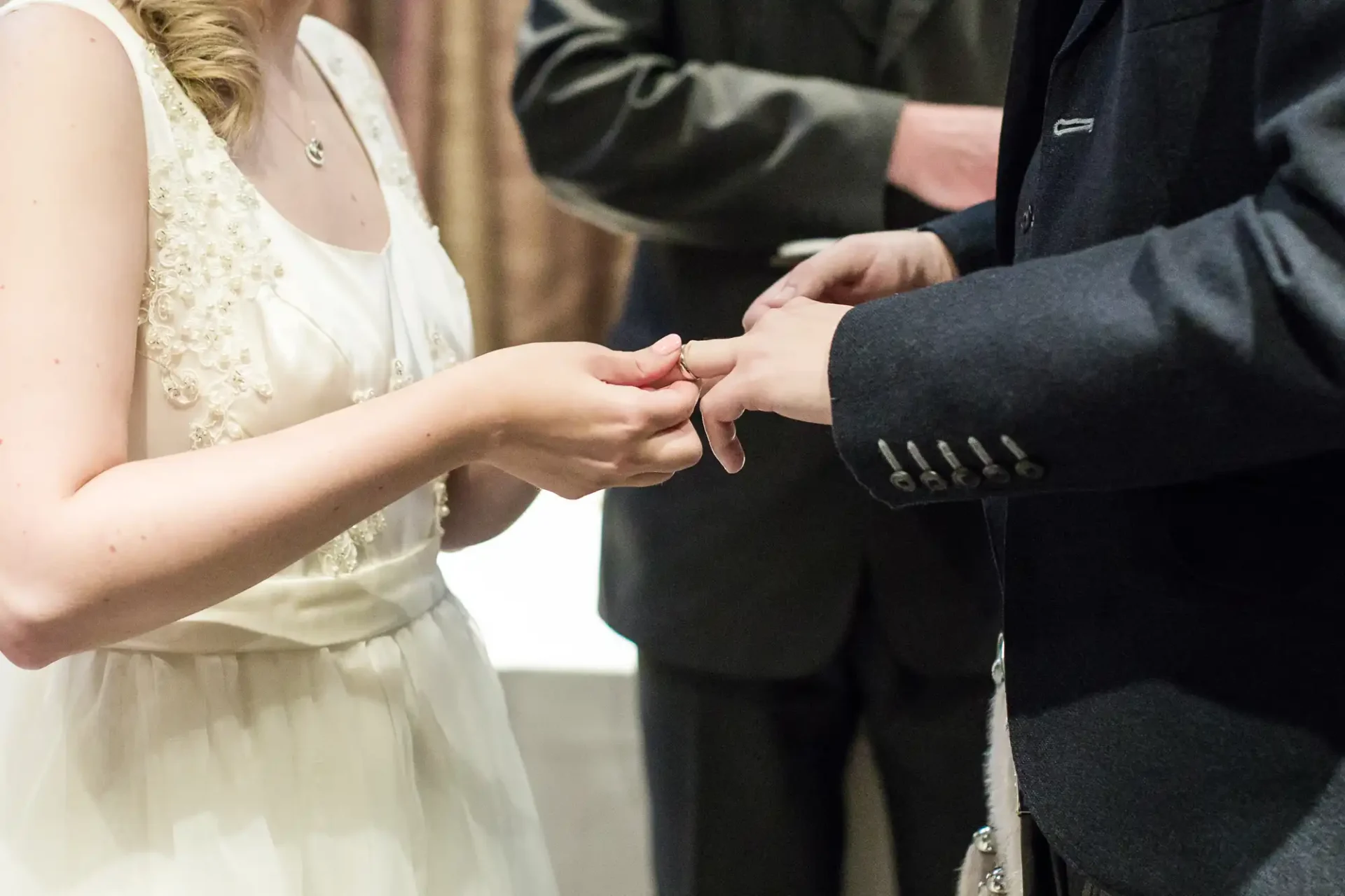 A bride placing a wedding ring on the groom's finger during a ceremony, with focus on their hands.