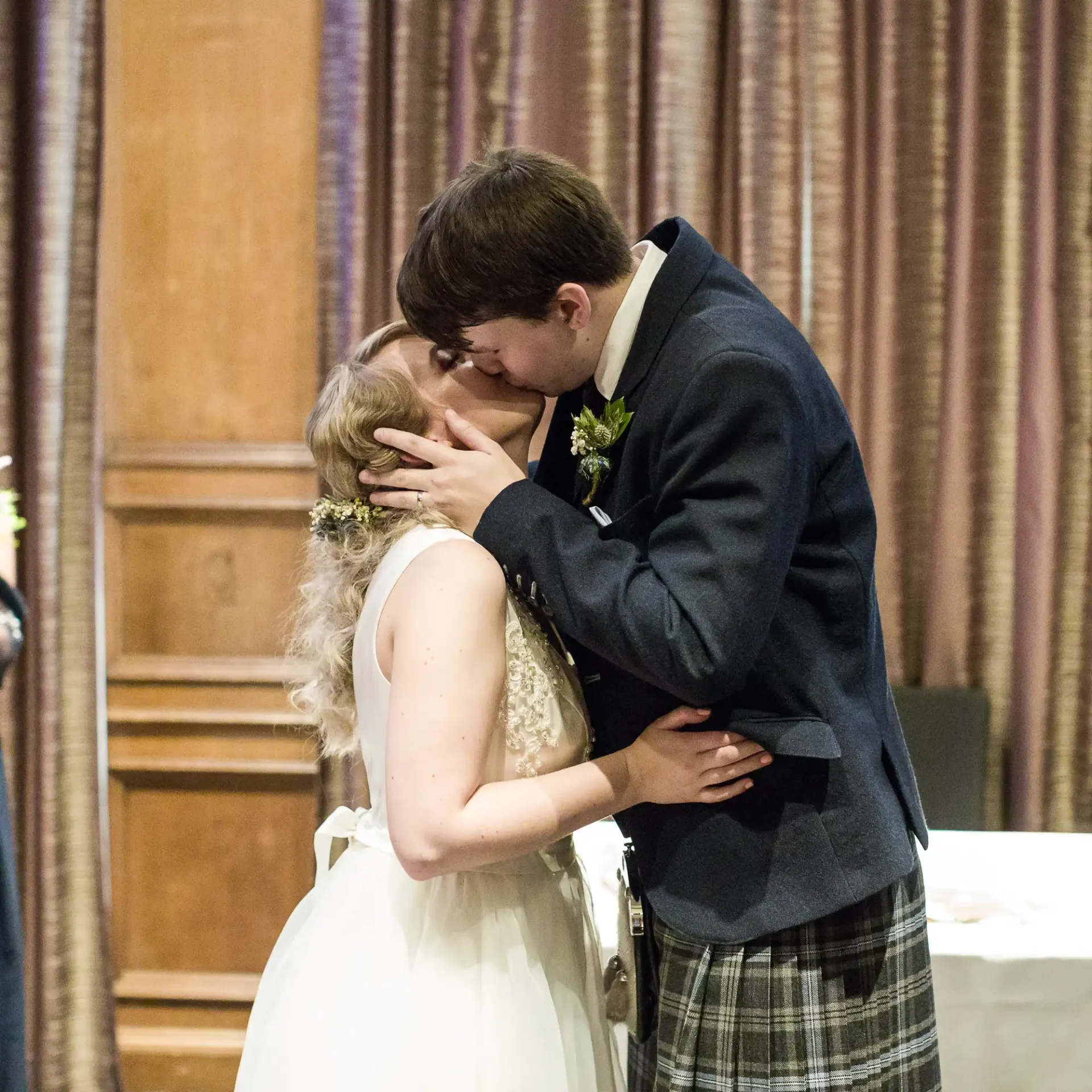 A bride and groom in wedding attire sharing a kiss, with the groom wearing a kilt and the bride a white dress with floral headpiece.