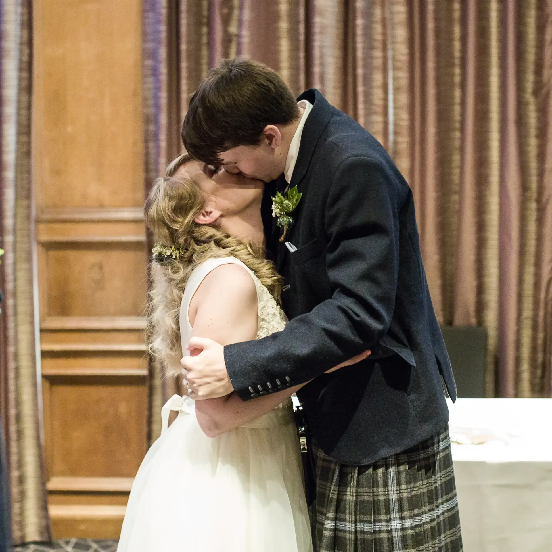 A bride and groom kissing passionately at their wedding reception, with the groom wearing a kilt and the bride in a white dress.