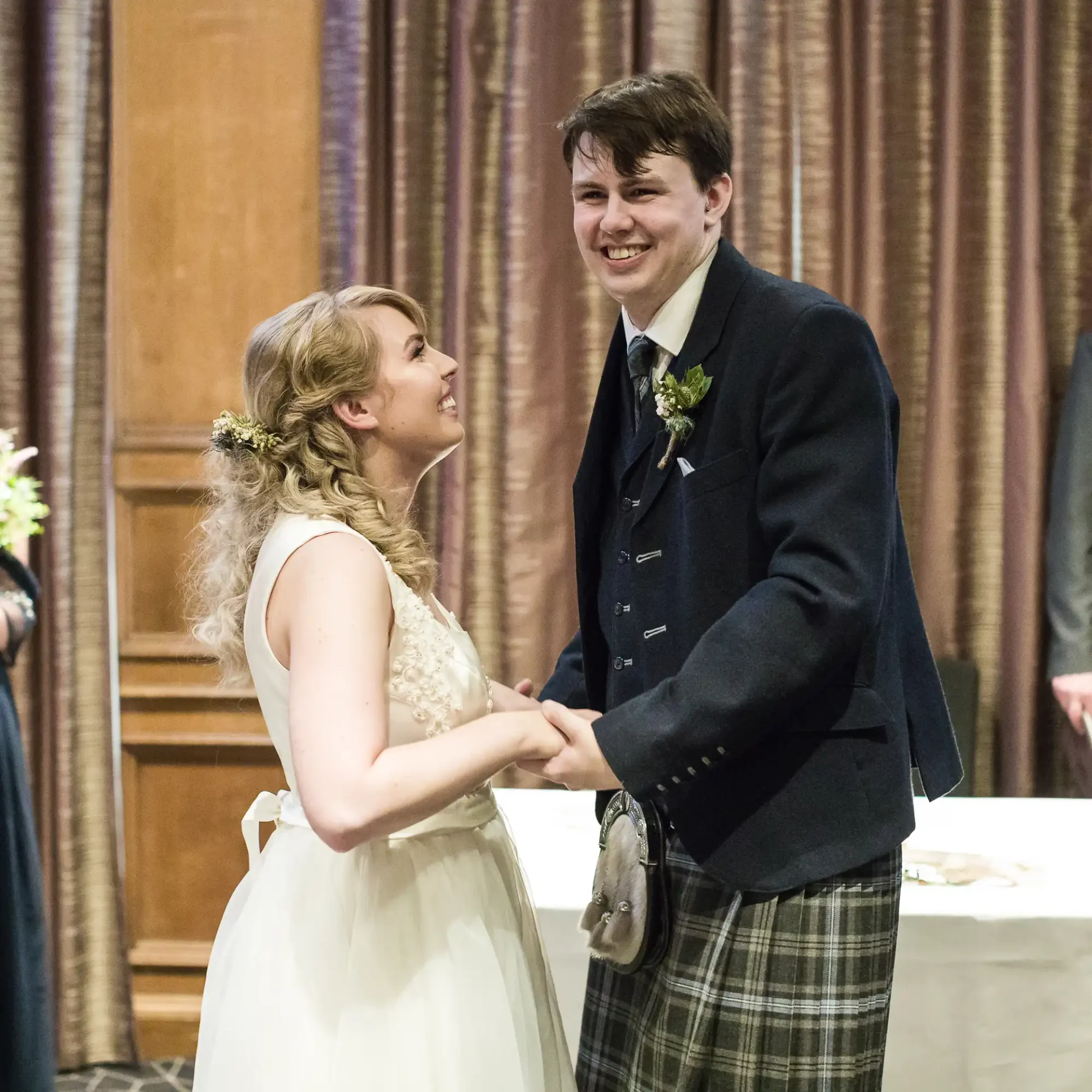 A bride and groom smiling at each other in a ceremonial hall, the groom in a kilt and the bride in a white dress with floral accessories in her hair.