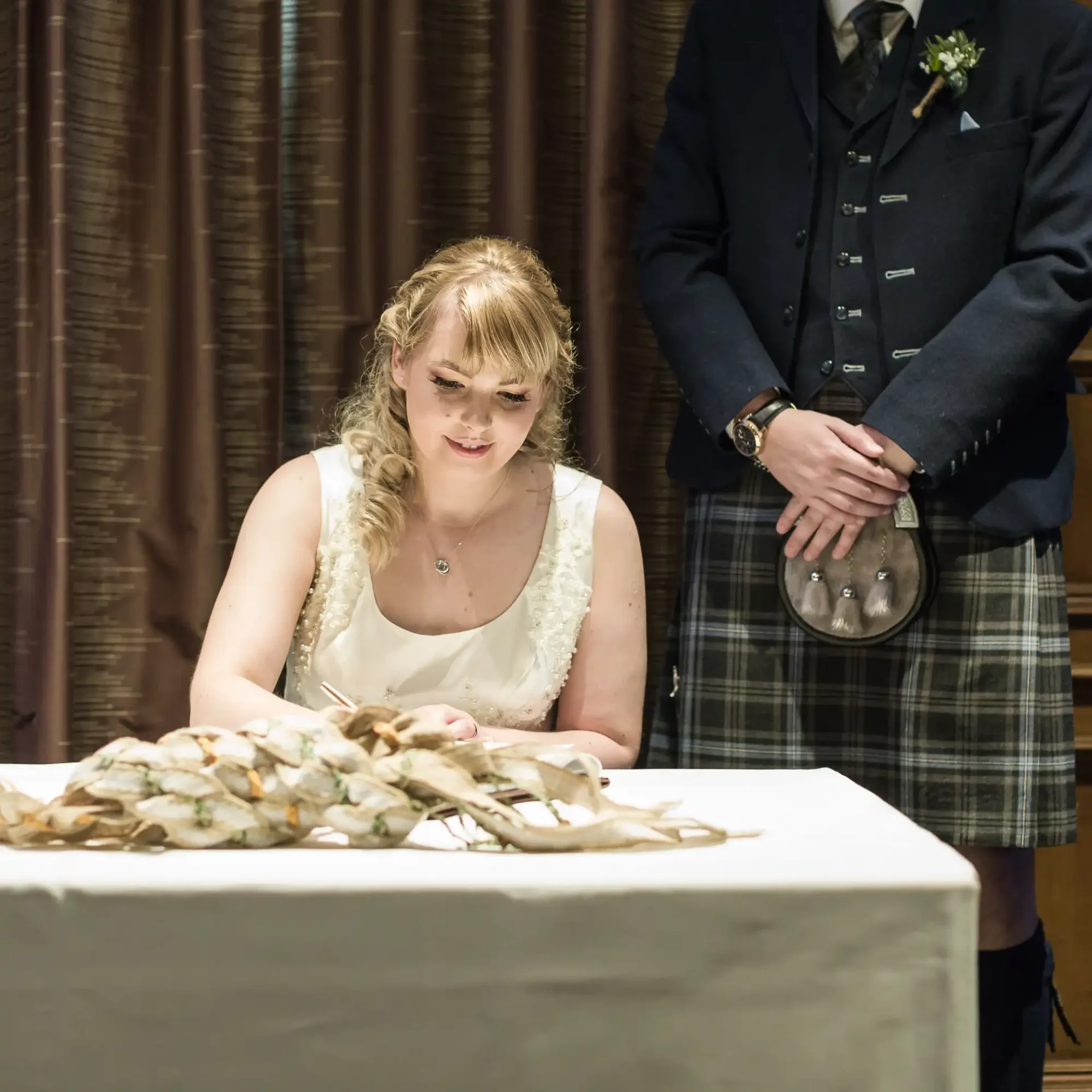 A woman in a white dress signs a document at a table with a bouquet in front of her, as a man in a kilt and suit jacket stands beside her.