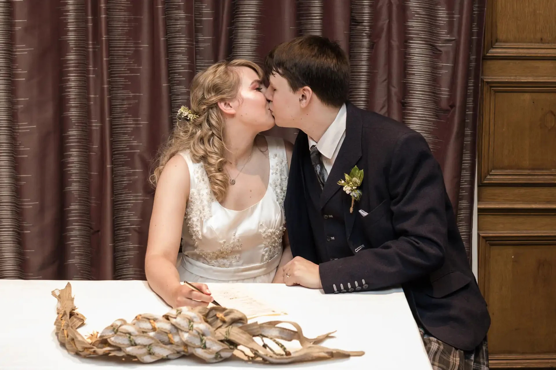 A bride and groom kissing at a signing table with a feather quill on it, in a room with wooden paneling and curtains.