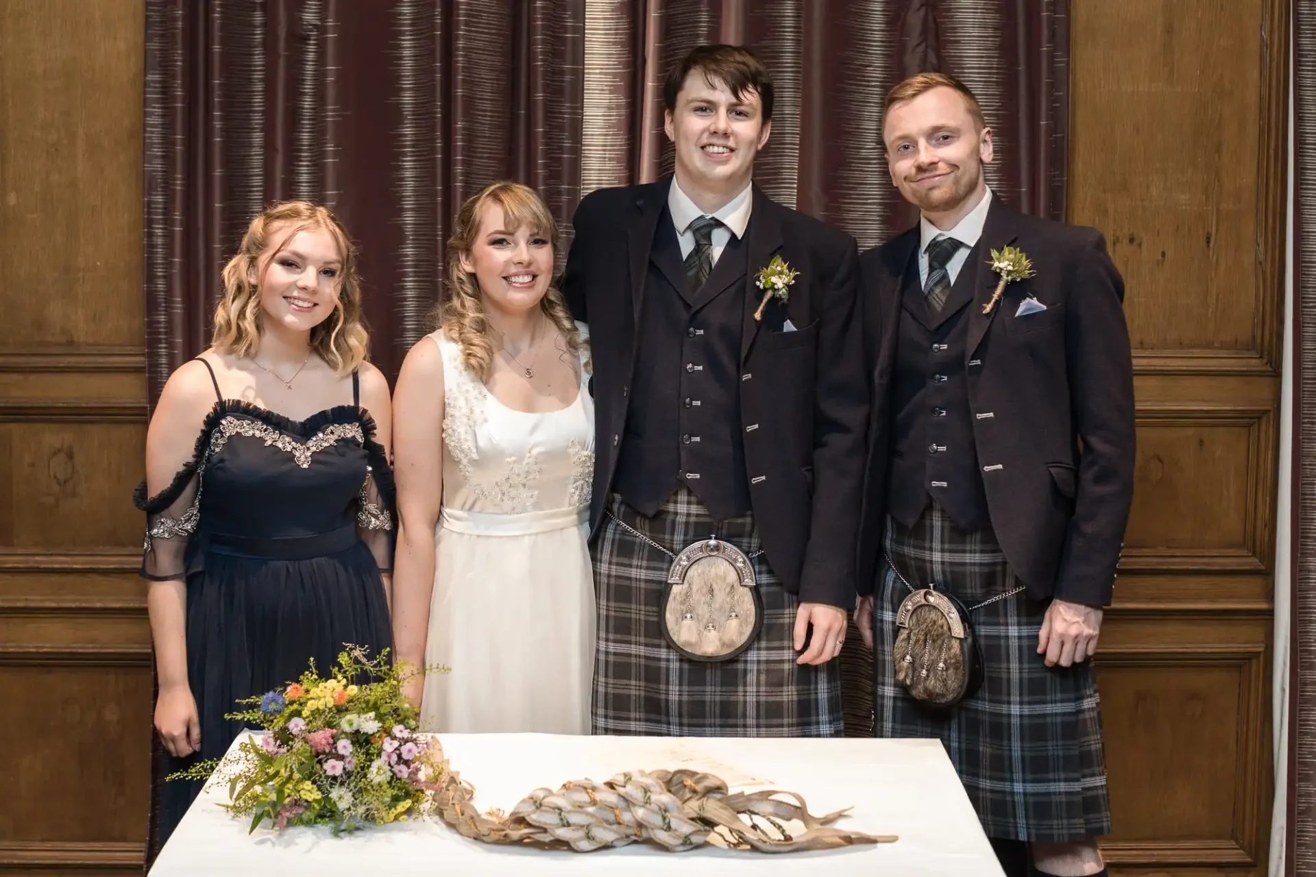 Two women and two men in formal attire, featuring kilts, posing with smiles behind a table adorned with flowers and antlers.