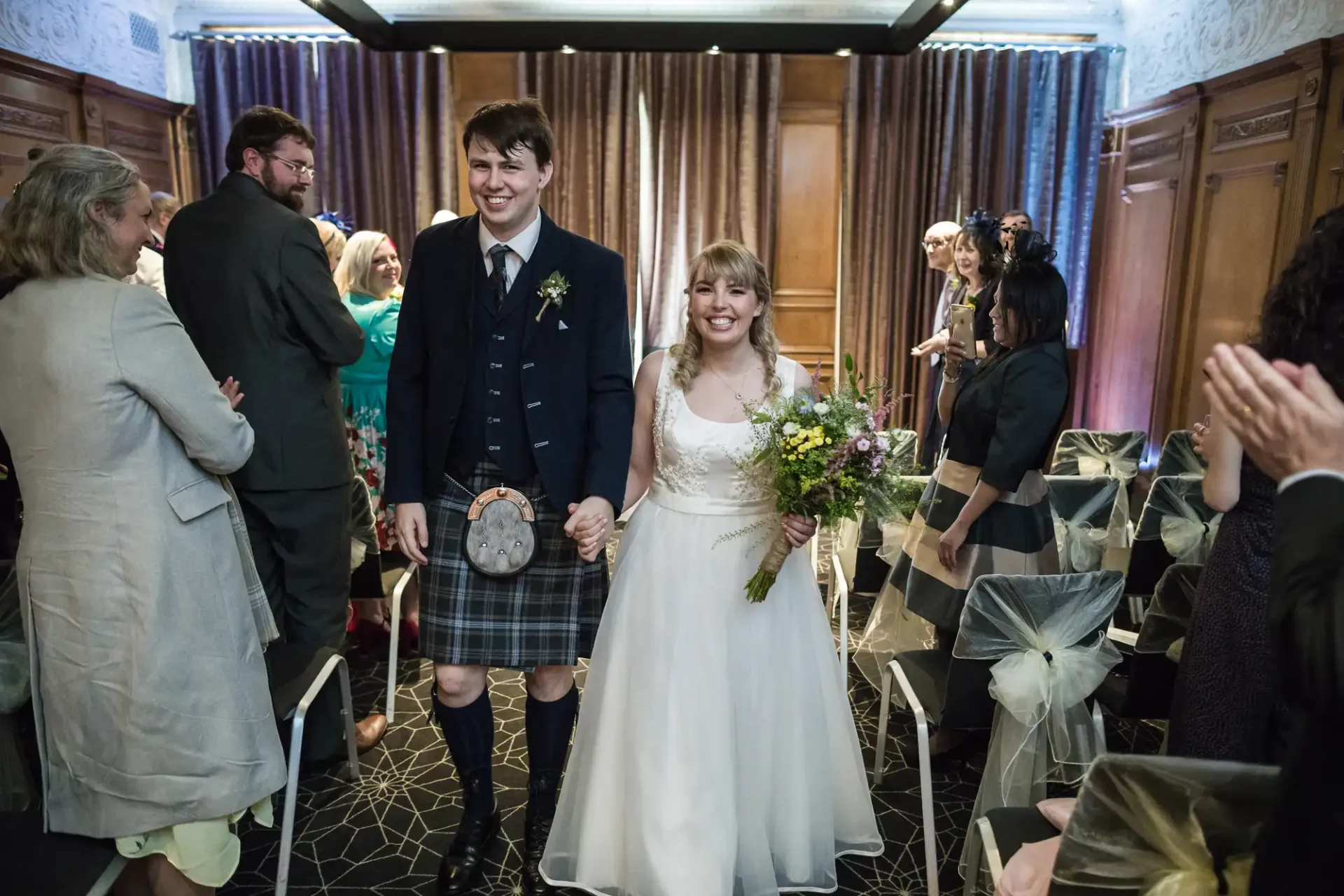 A smiling bride and groom in traditional scottish attire walk down the aisle, surrounded by applauding guests in an elegantly decorated hall.