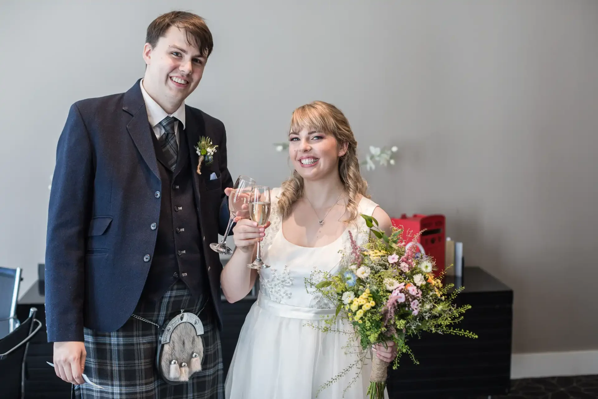 A smiling bride and groom in wedding attire, the groom in a kilt, holding a glass and a bouquet, standing indoors.