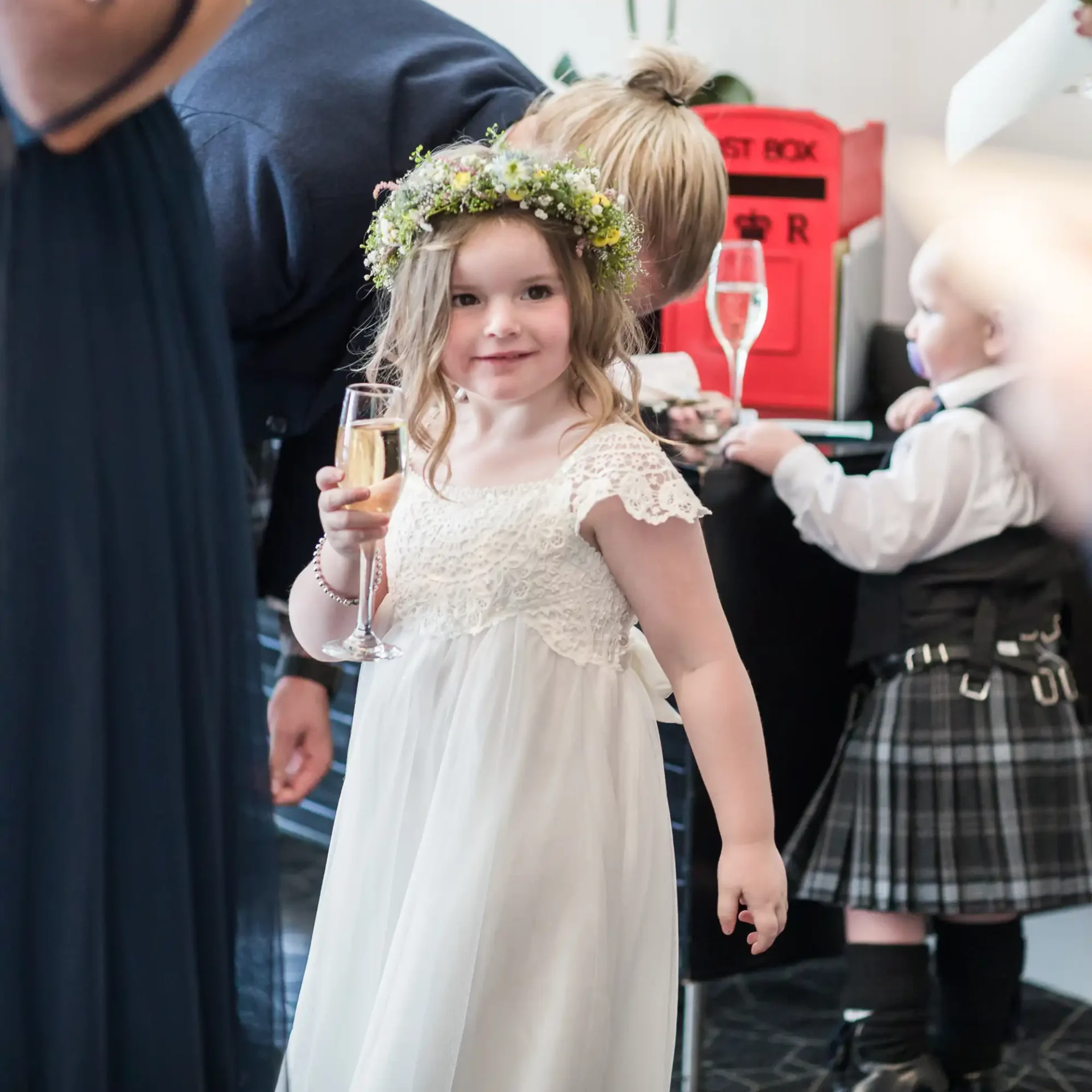 A young girl in a white dress and floral wreath smiles while holding a champagne glass at a festive gathering.