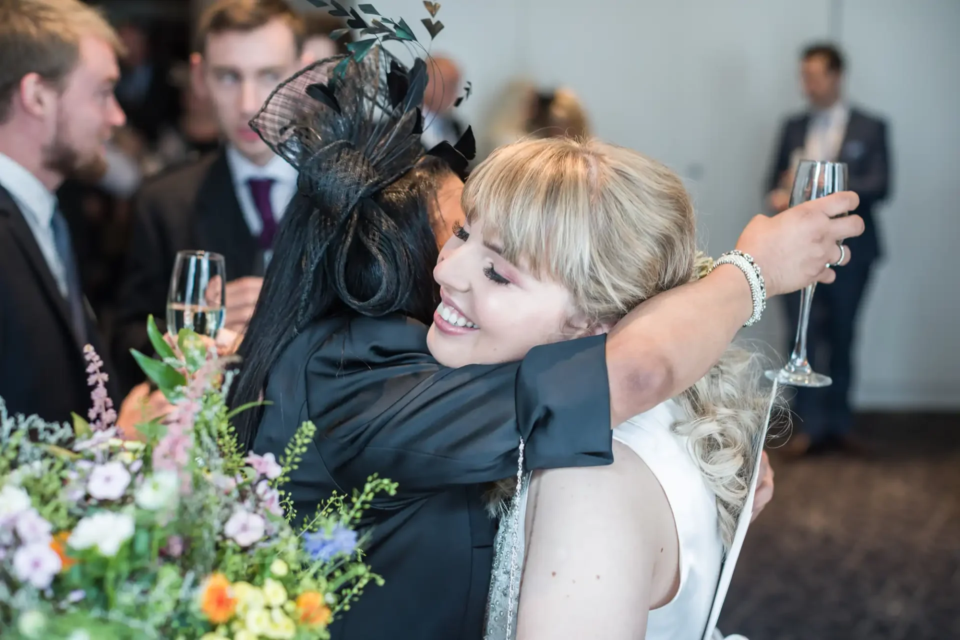 A joyful bride in a white dress hugs a guest wearing a black outfit and a large hat at a wedding reception, with other guests nearby holding champagne glasses.