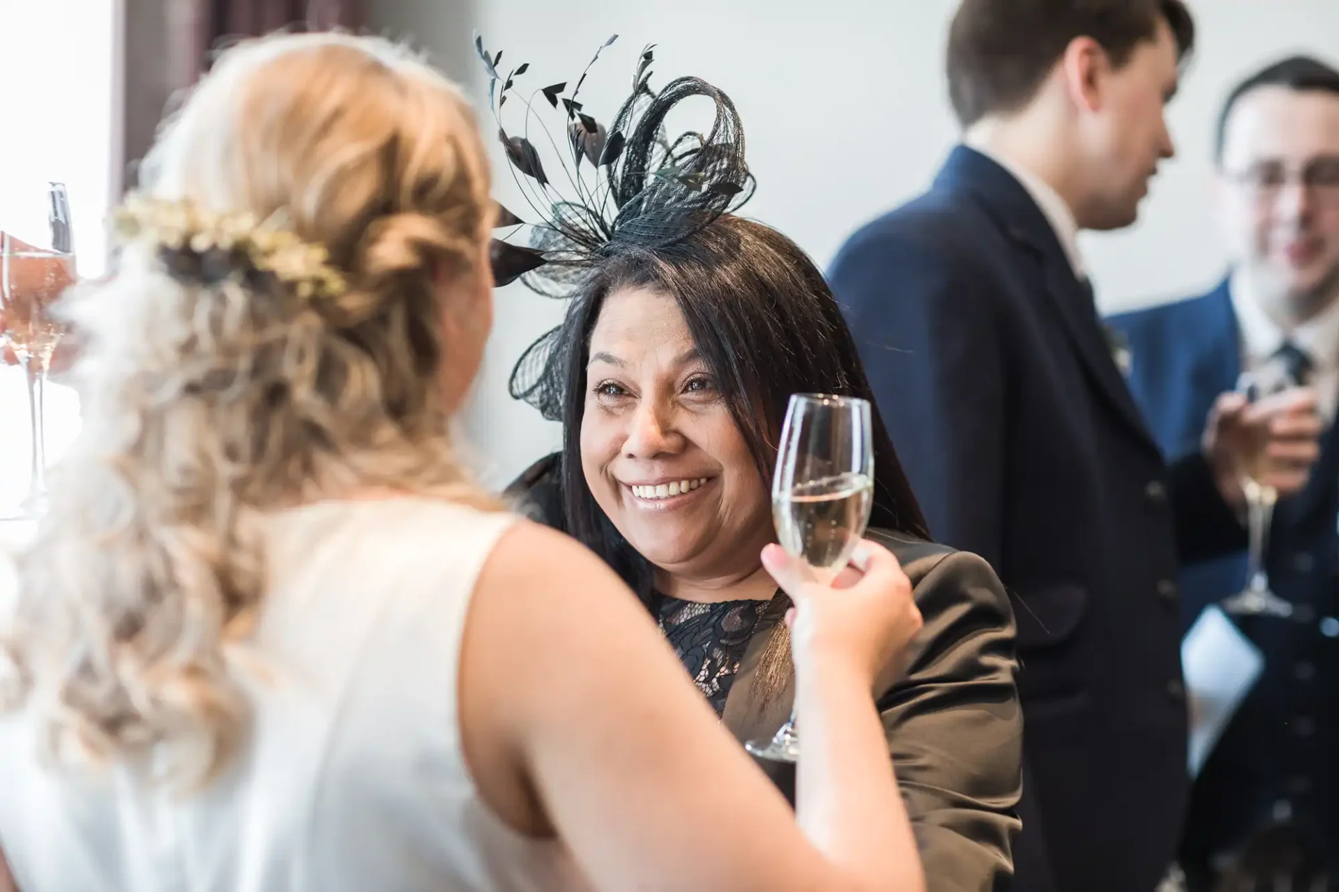 A woman in a black dress with a feathered headpiece smiles while holding a champagne glass, conversing with another guest at a social gathering.