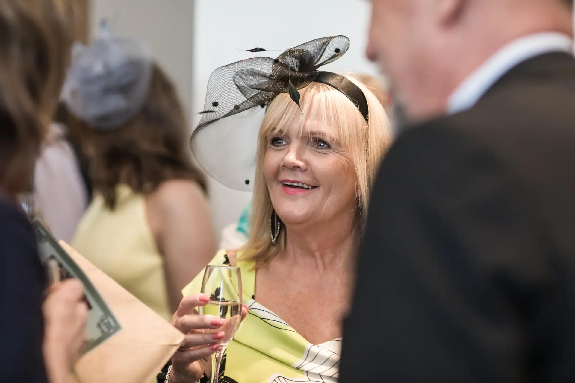 A woman in a black and white hat holding a champagne glass converses at a social event.
