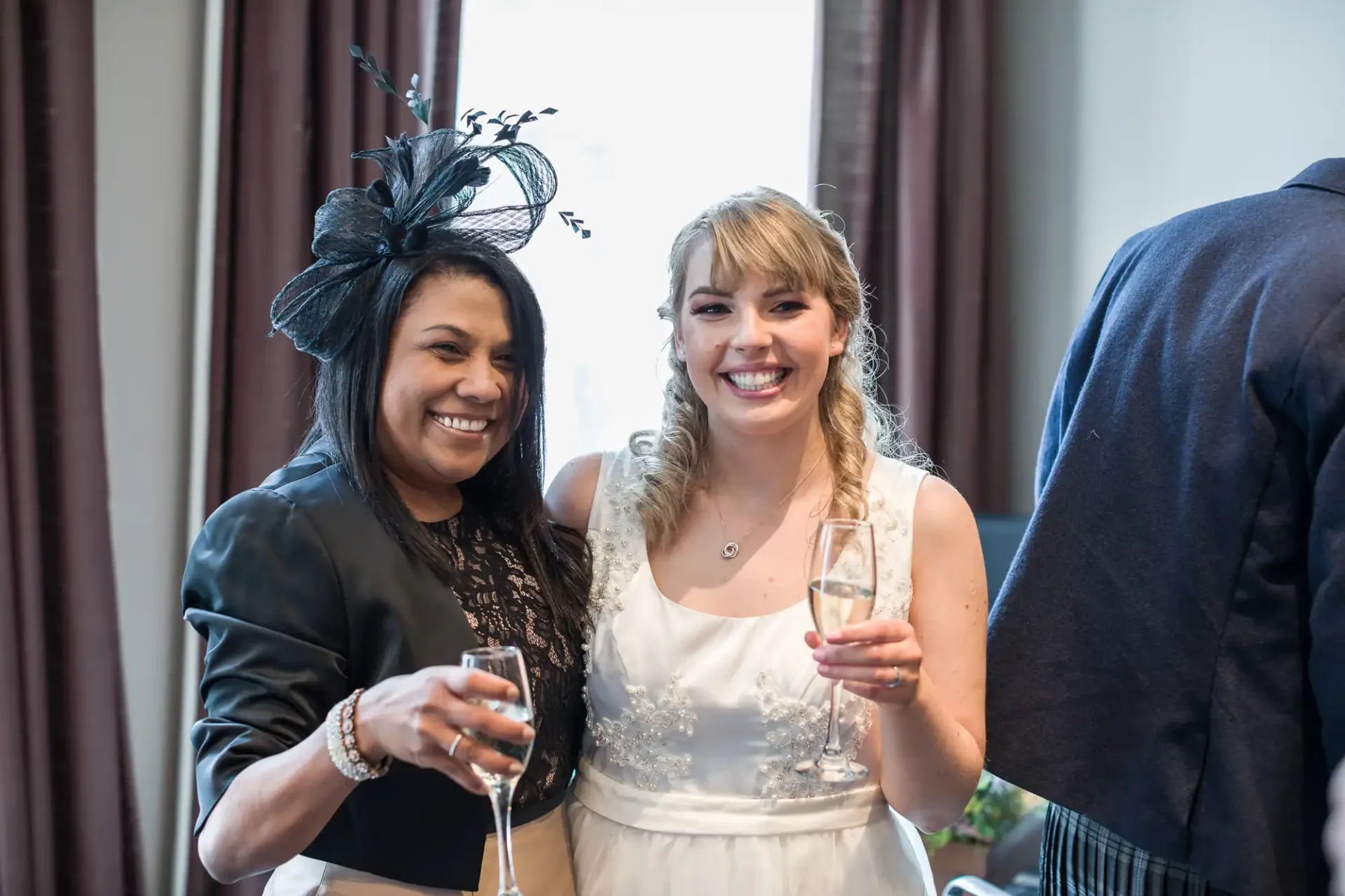 Two women smiling and toasting with champagne glasses at a celebration, one in a black outfit with a hat and the other in a white bridal dress.