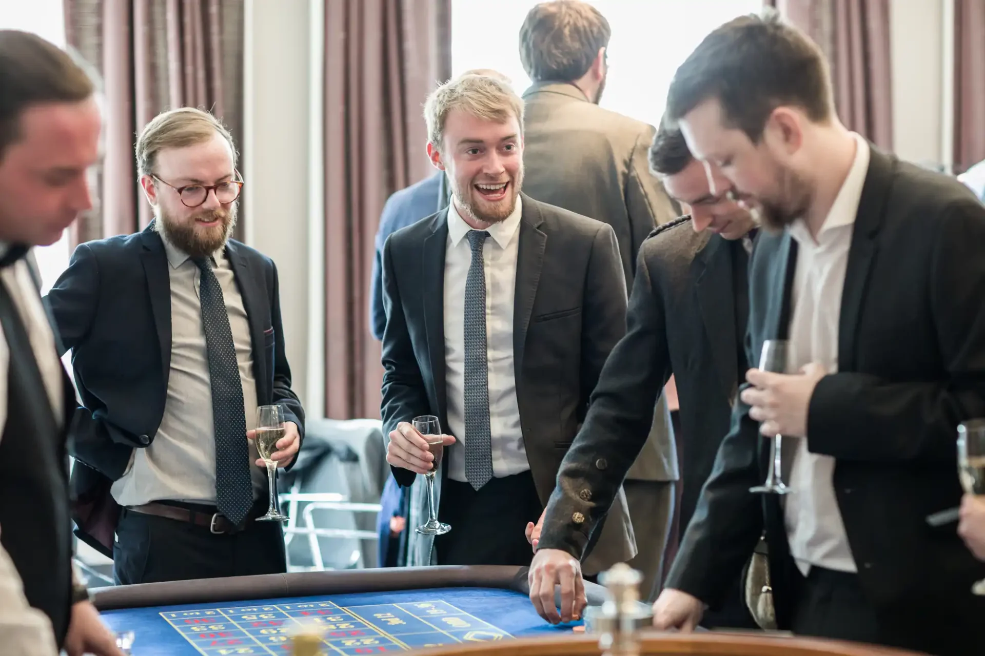Group of men in suits laughing and engaging in a game at a casino table, holding wine glasses.