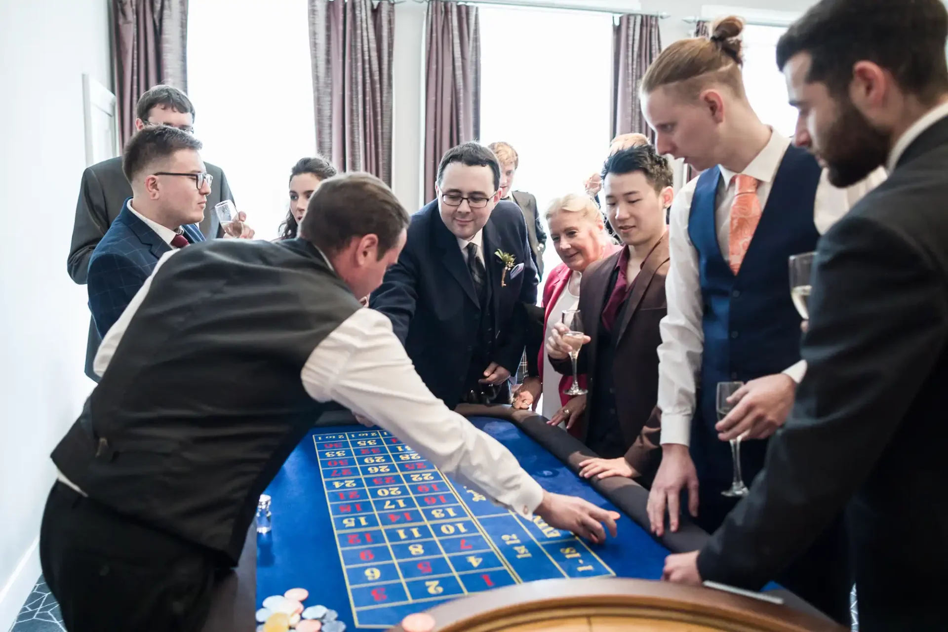 A group of people, including a man in a boutonniere, watching a dealer at a roulette table during an indoor event.