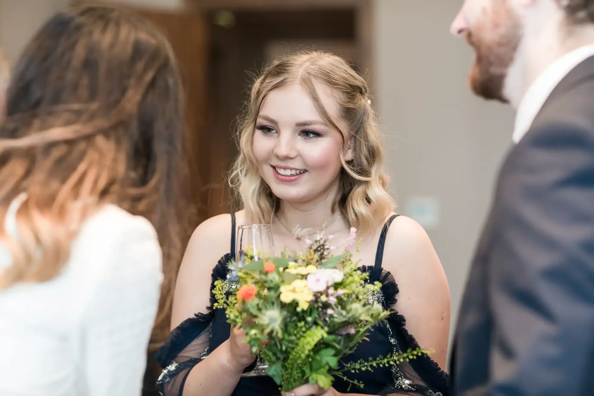 A woman in a black dress holding a bouquet converses with guests at a social event.