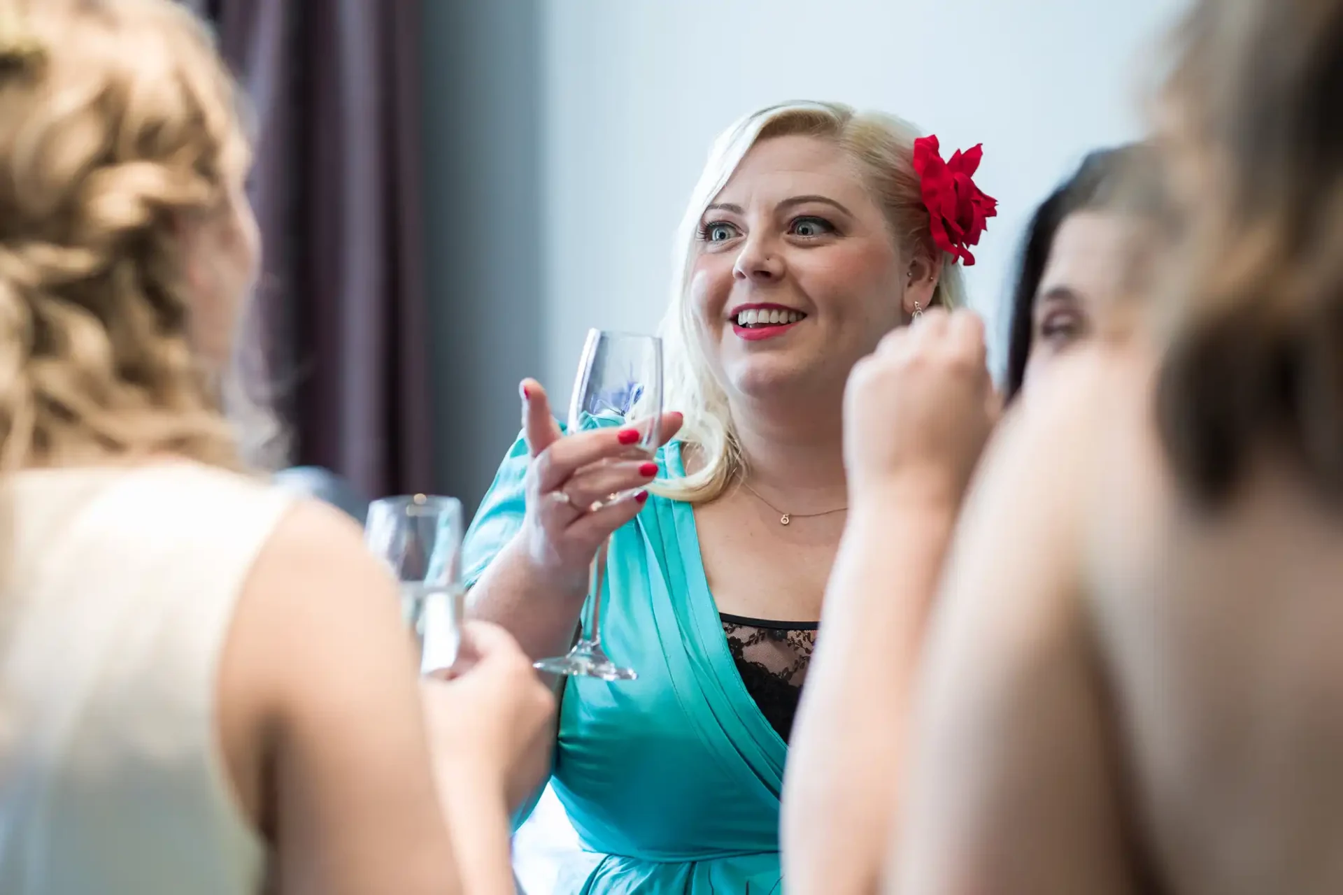 A woman in a teal dress with a red flower in her hair smiling and holding a glass of wine at a social gathering.