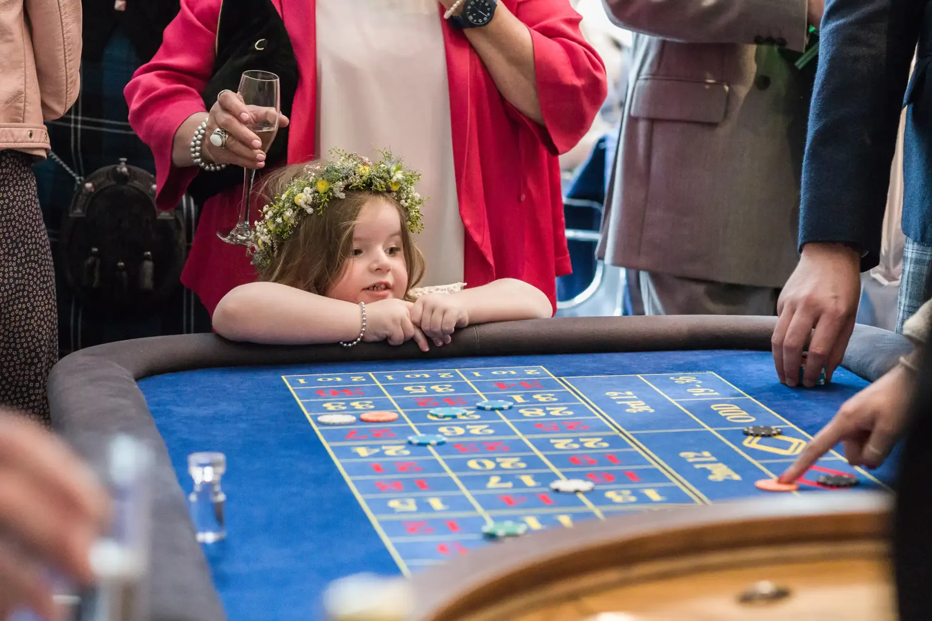 A young girl wearing a floral wreath on her head leans on a casino table, watching a roulette game in progress, surrounded by adults.