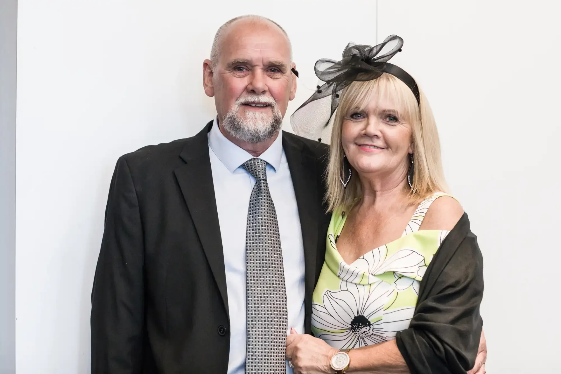An older man and woman posing together; he is in a suit and she wears a floral dress with a black fascinator.