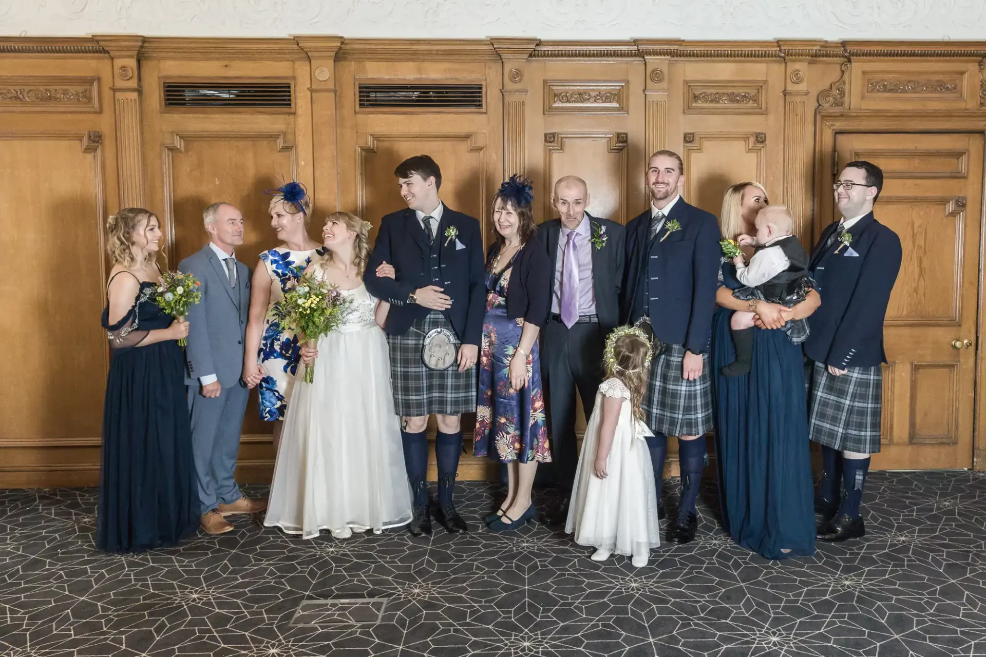 A family dressed in formal attire, including kilts, posing in a line at a wedding event, with a variety of happy expressions and interactions.