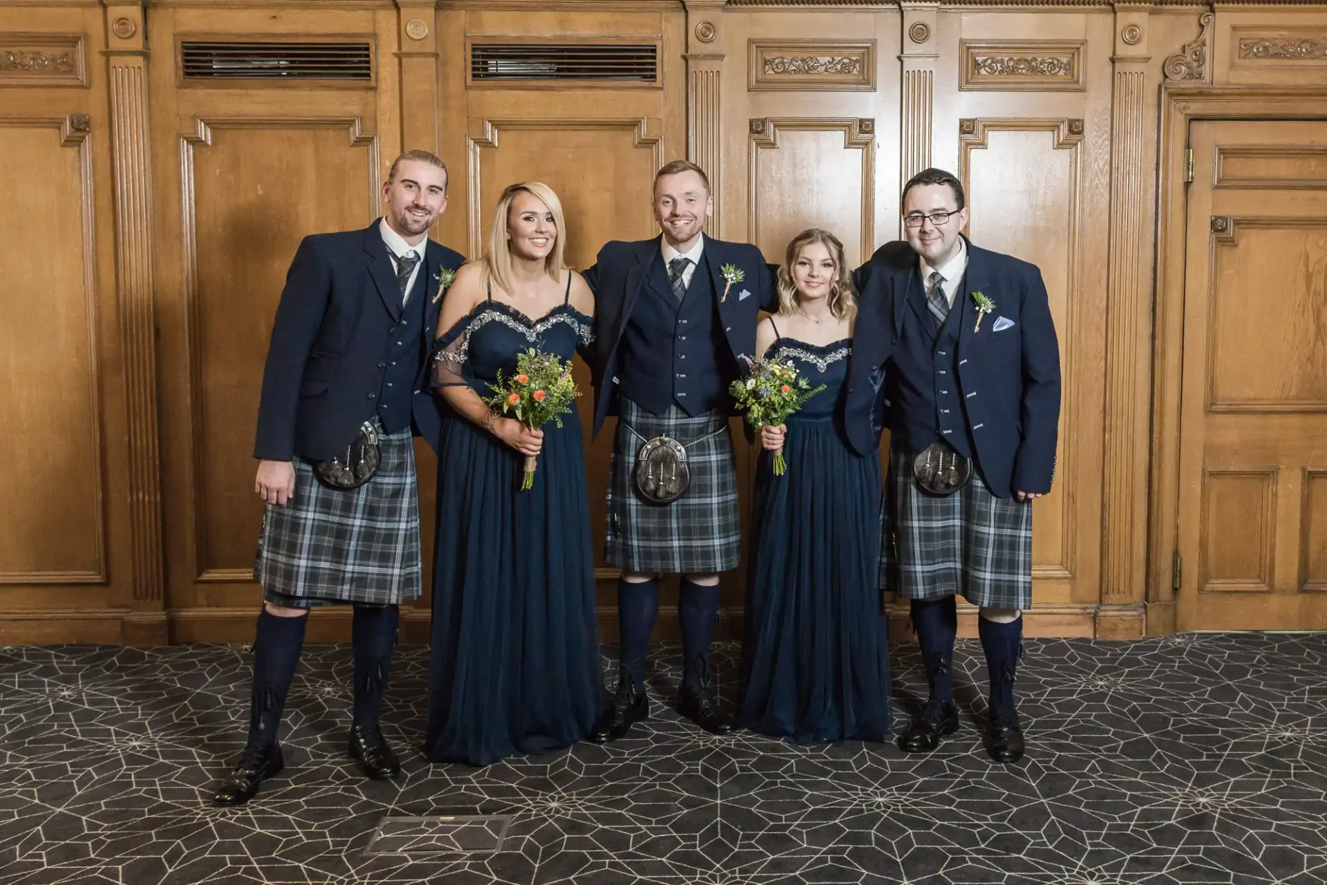 Five people in formal attire with kilts and dresses posing together at a wedding in a wood-paneled hall.