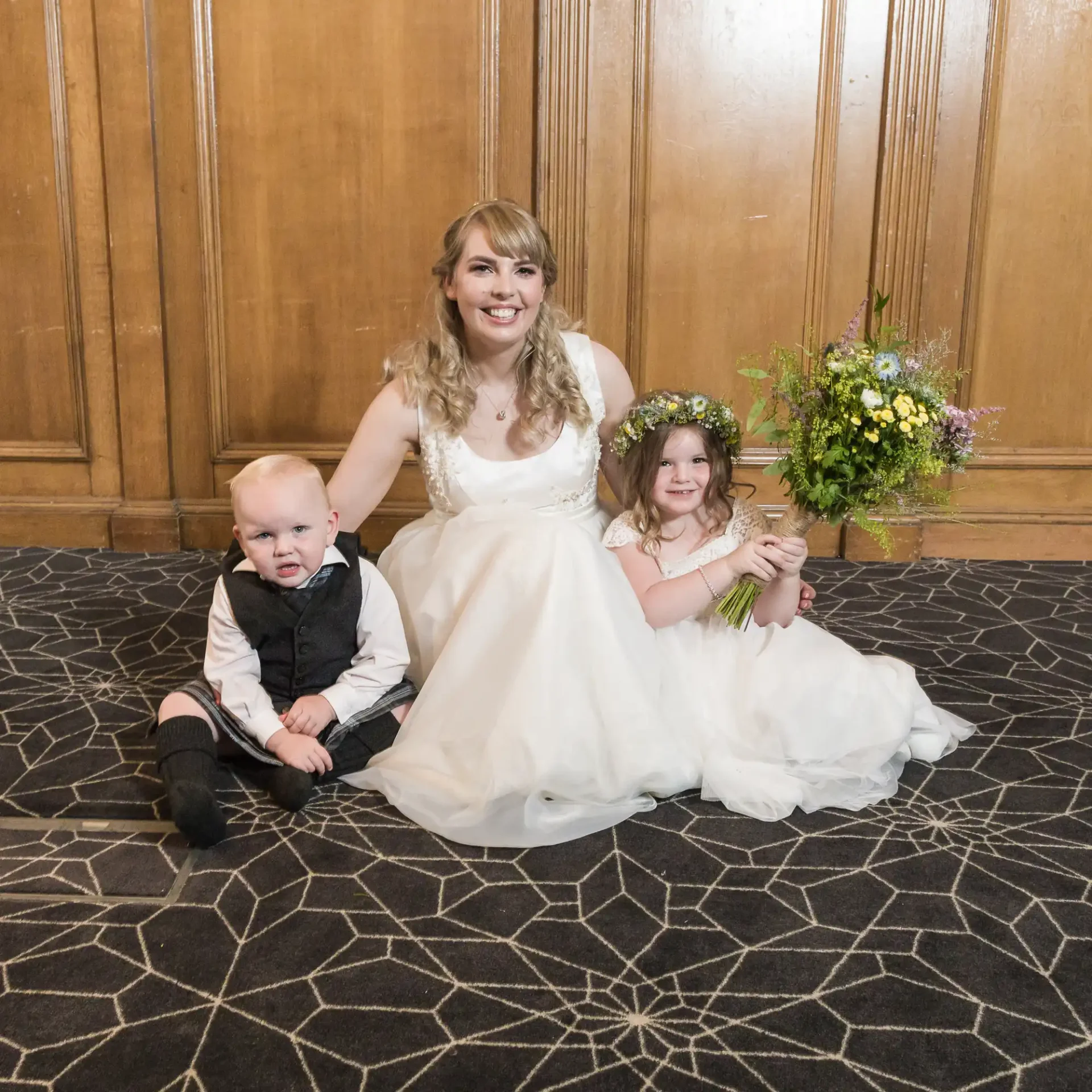 A bride in a white dress sits on the floor with a young boy in a black kilt and a little girl in a white dress, all smiling, in front of a wooden panel and floral bouquet.
