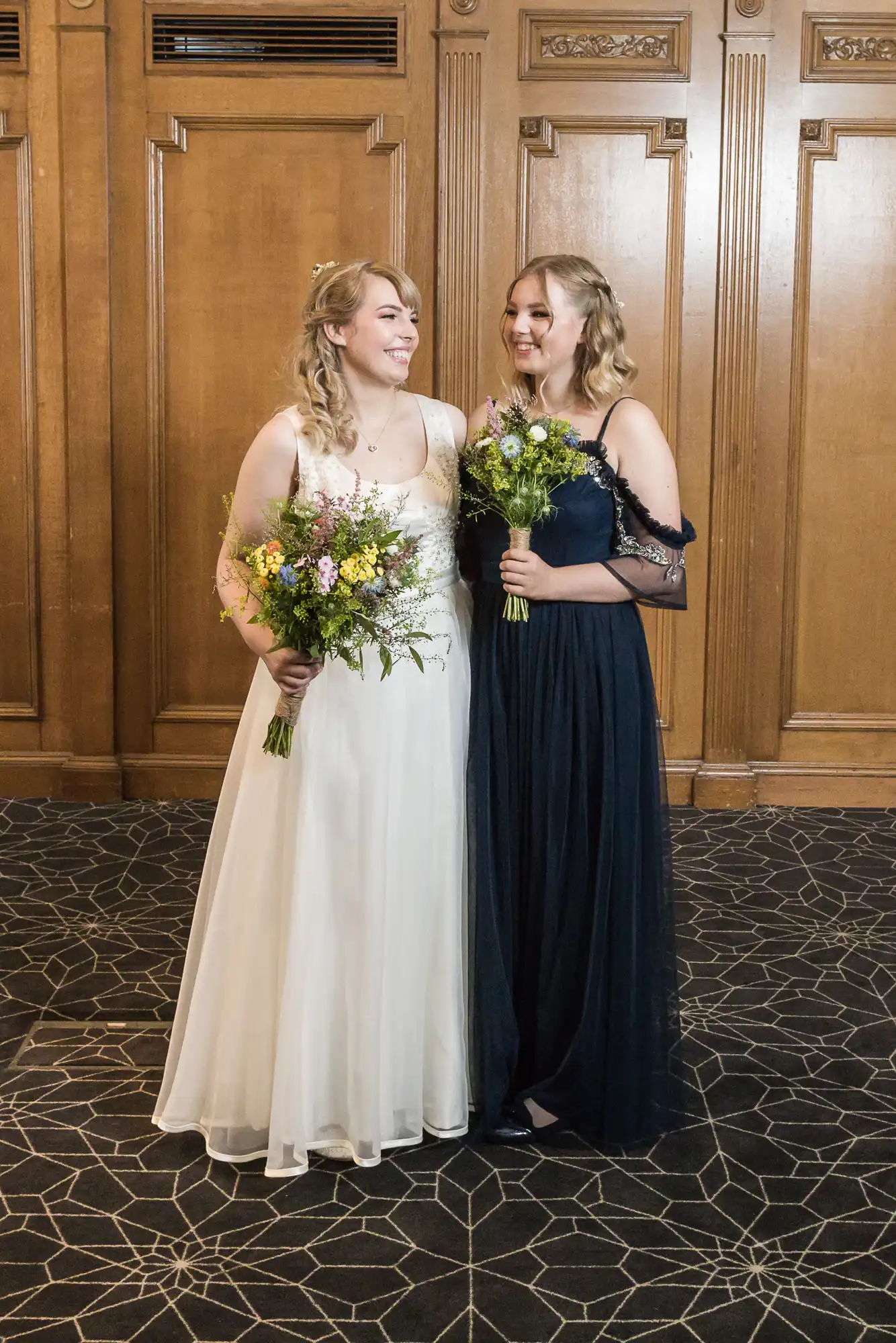Two women in elegant dresses, one in white and one in navy blue, smiling and holding bouquets in a grand hall.