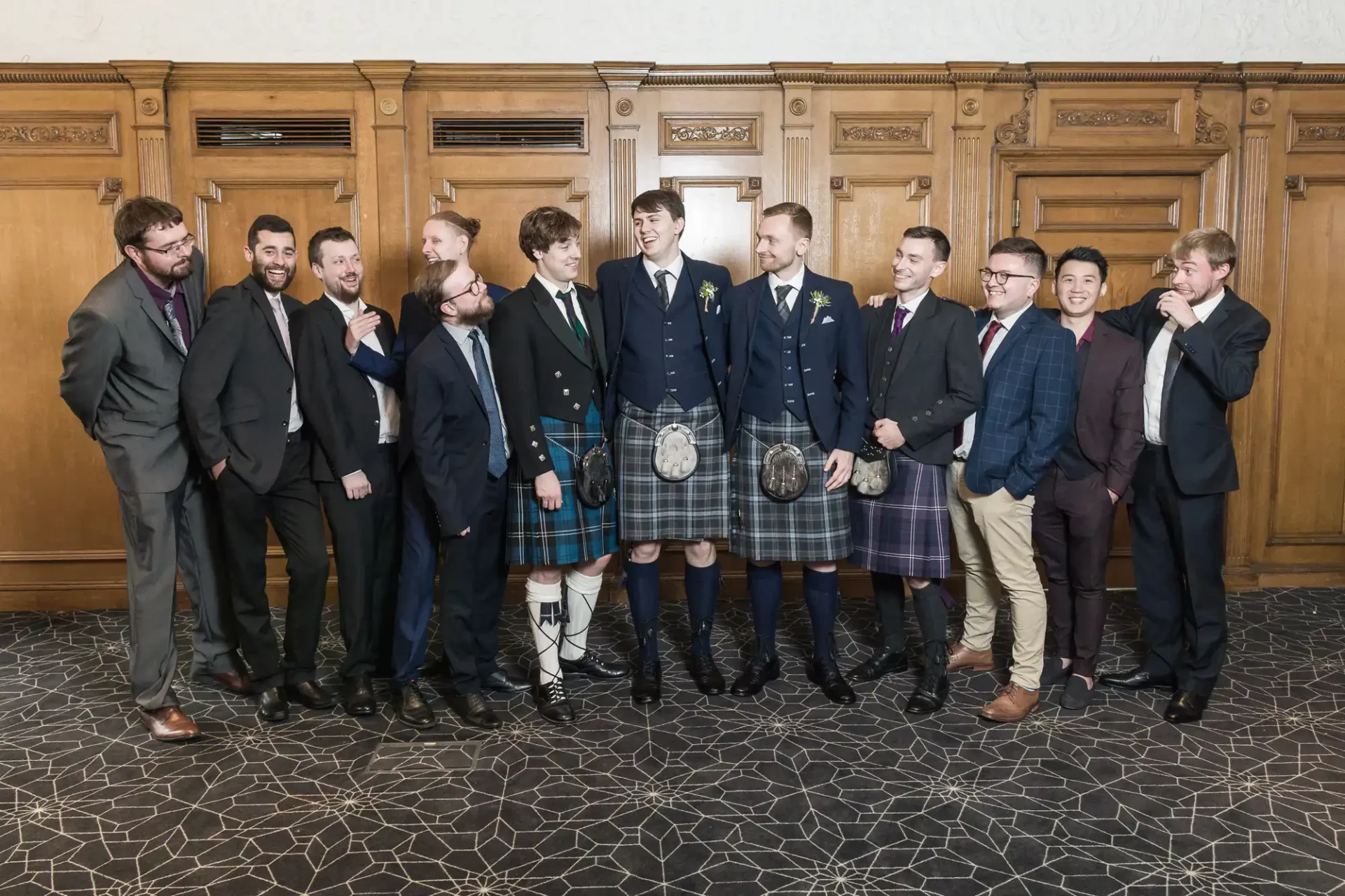 Group of men in formal attire, including suits and traditional kilts, smiling and posing together in a decorated room.