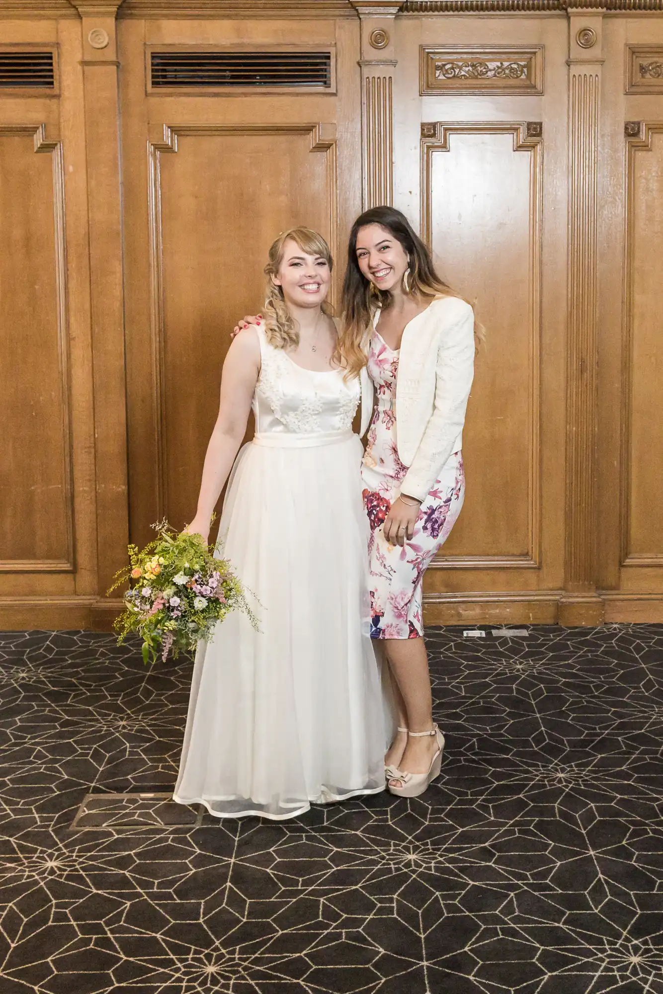 Two women smiling in a formal setting, one in a white wedding dress holding a bouquet and the other in a floral dress with a blazer.